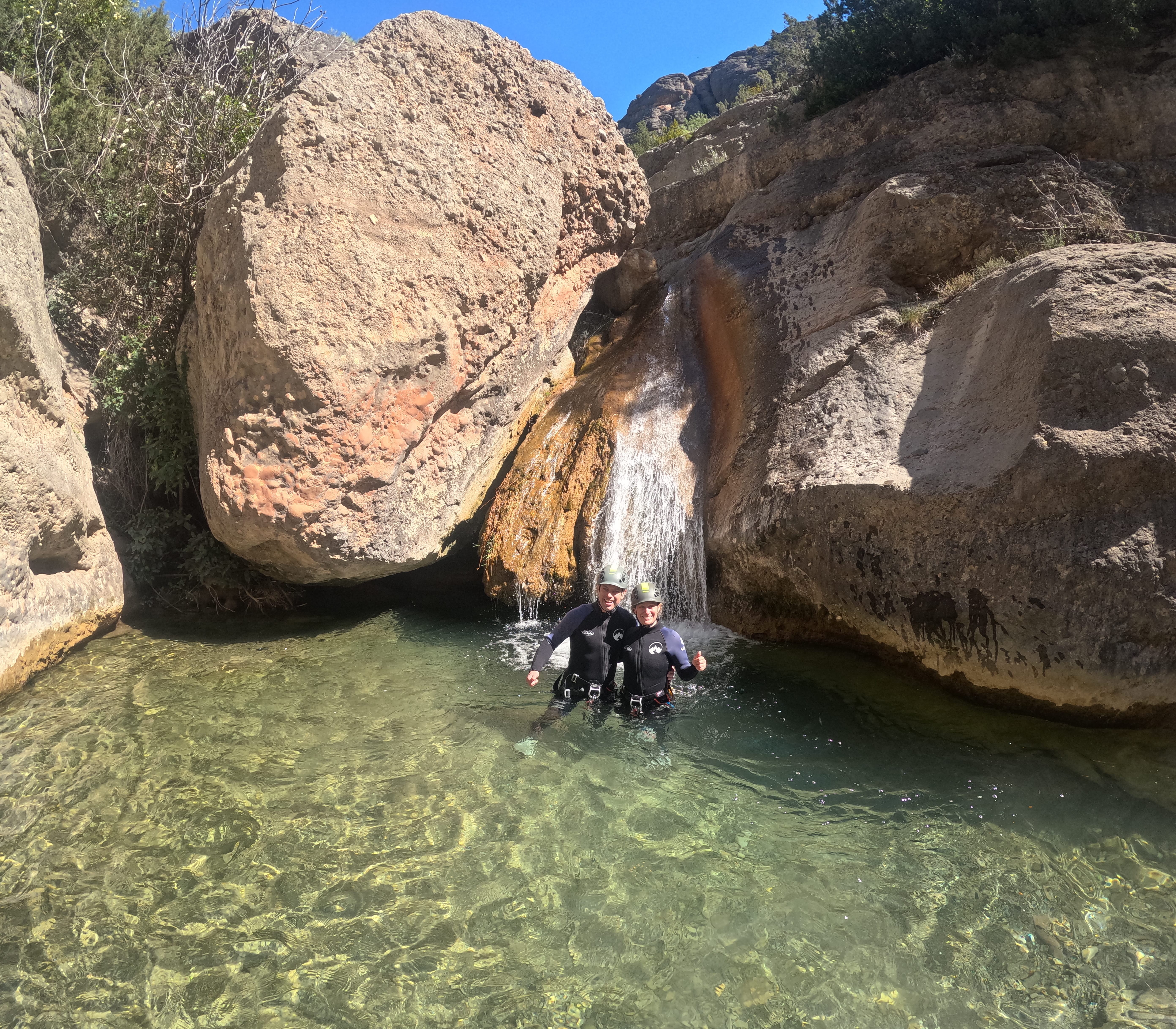 Two participants standing in a clear natural pool beneath a small waterfall surrounded by rocky walls in Barranco Sant Pere