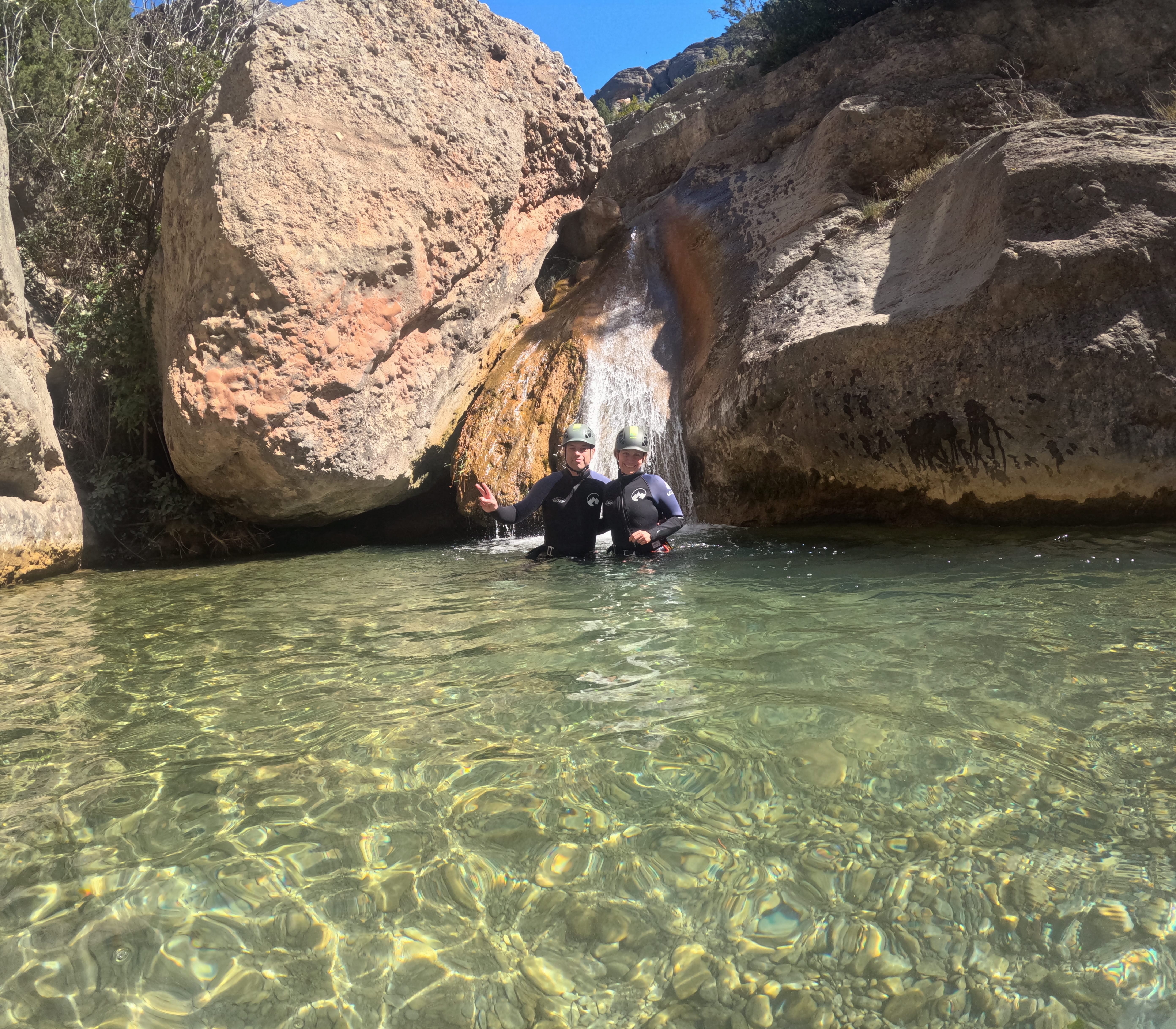 Two canyoning participants standing in a crystal-clear natural pool beneath a small waterfall in Barranco Sant Pere