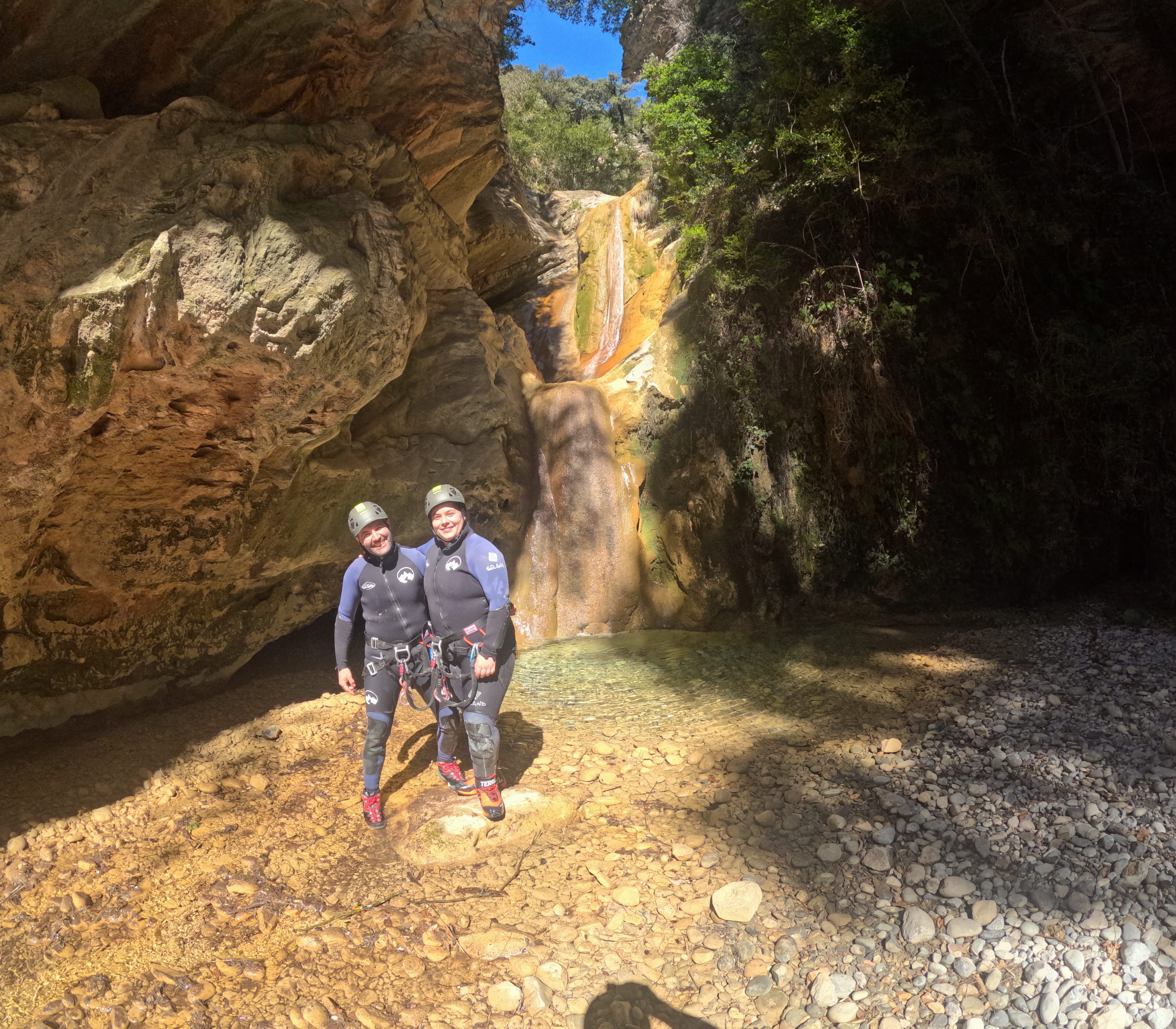 Two participants in canyoning gear posing beside a small waterfall and natural pool in Barranco de Gurp