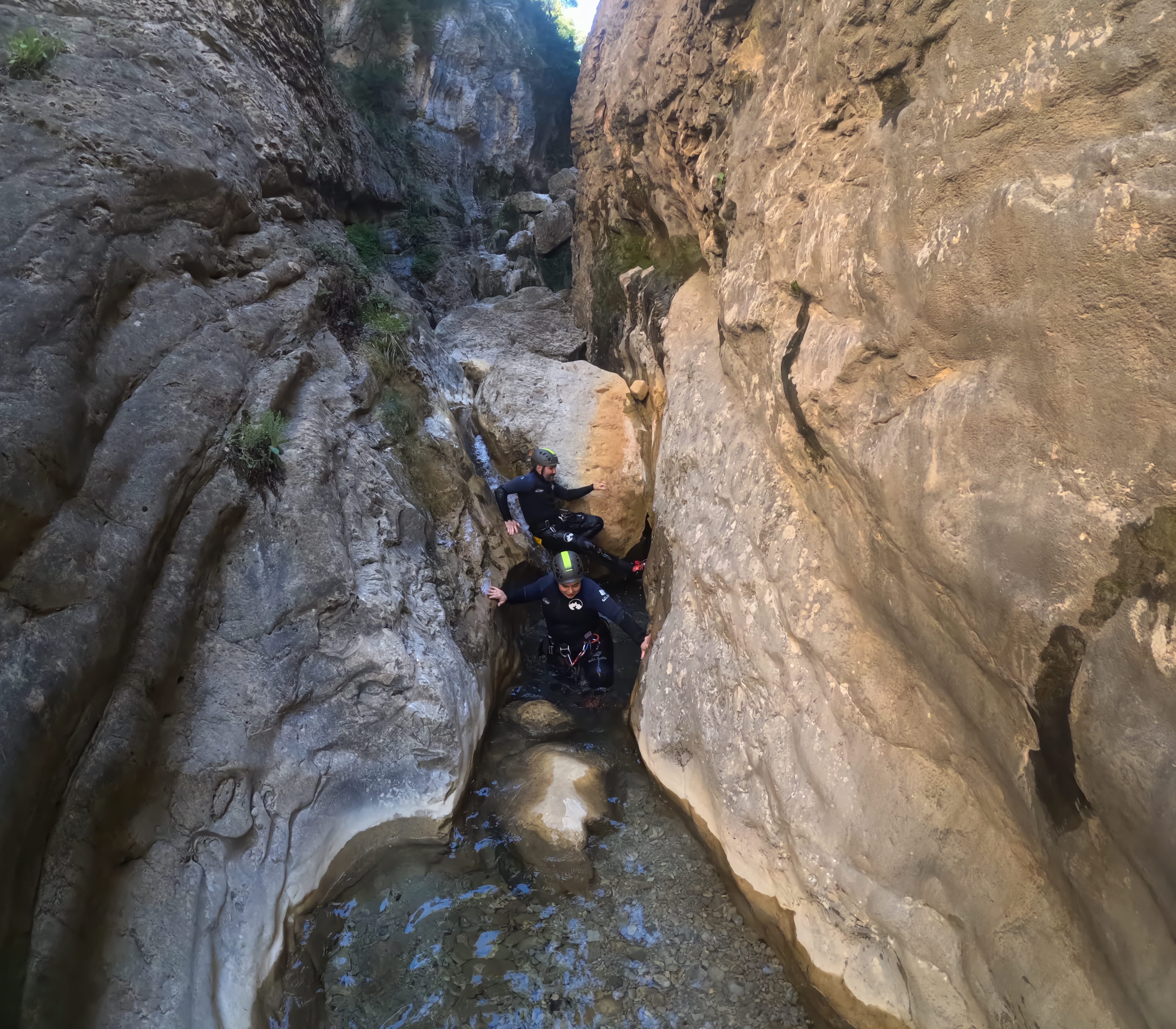 Two participants moving through a narrow rocky passage with shallow clear water in Barranco de Gurp