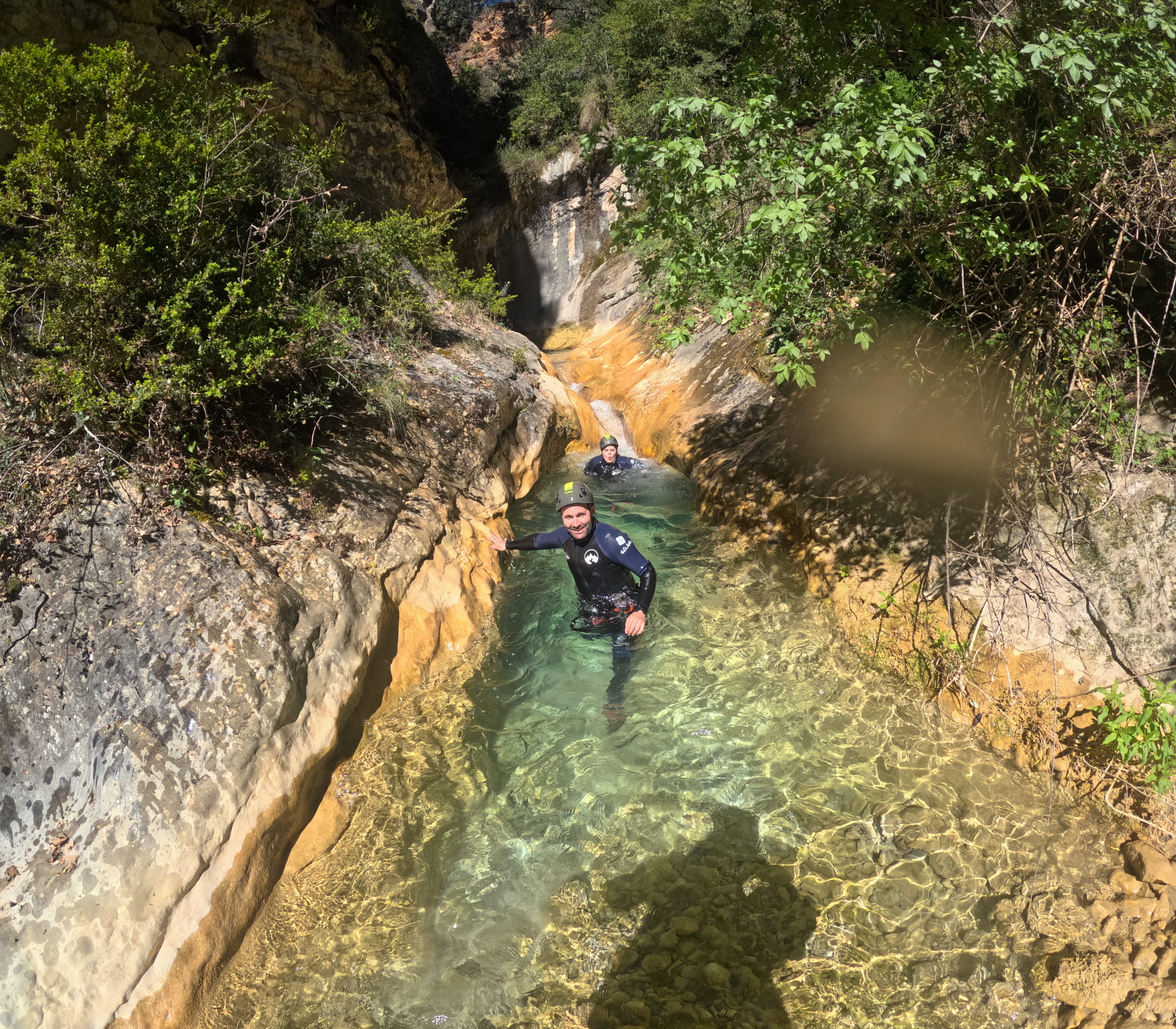 Two participants wading through a narrow crystal-clear natural pool surrounded by rocky walls and vegetation in Barranco de Gurp