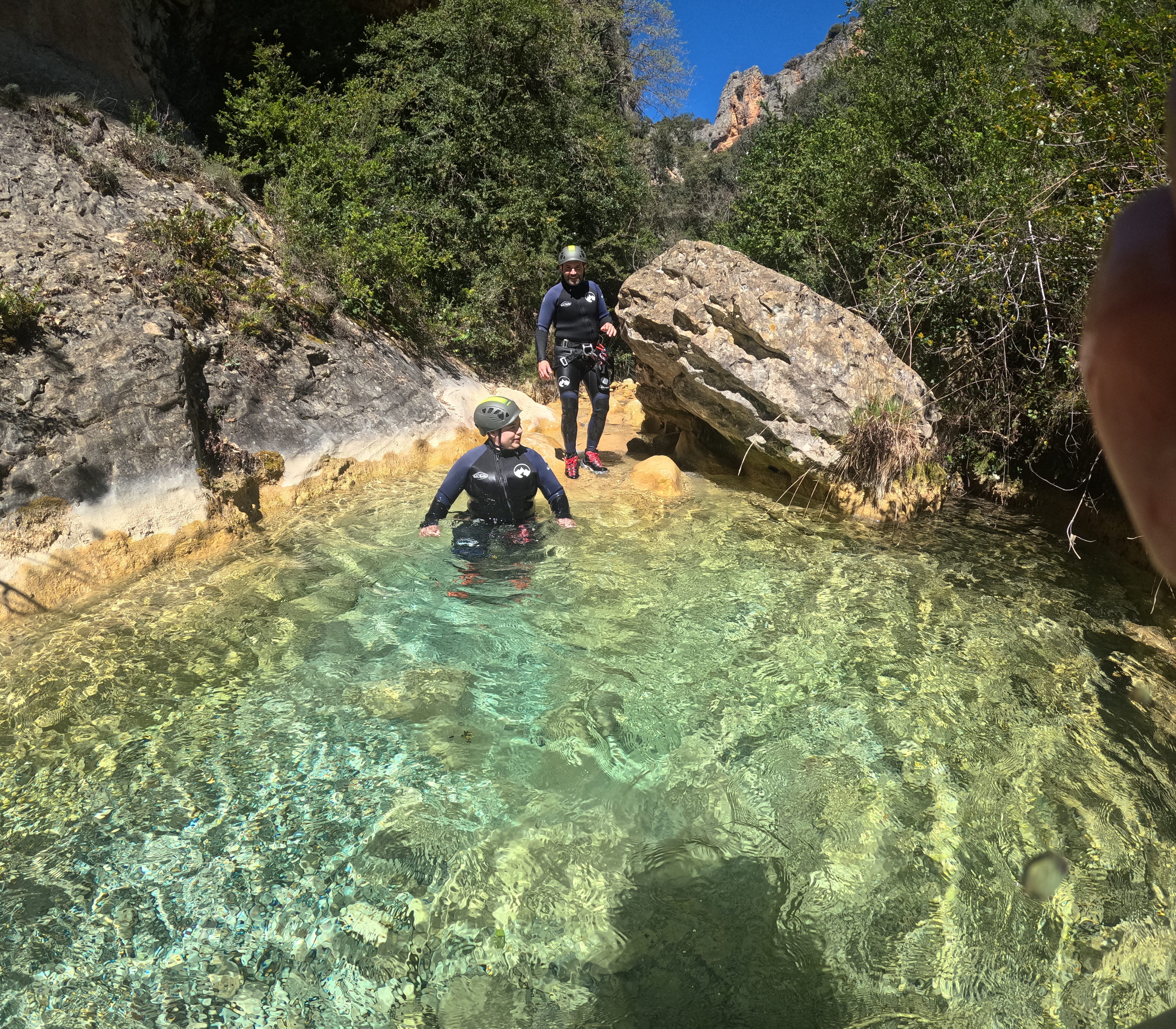 Two participants in canyoning gear crossing a clear turquoise natural pool among rocks and vegetation in Barranco de Gurp