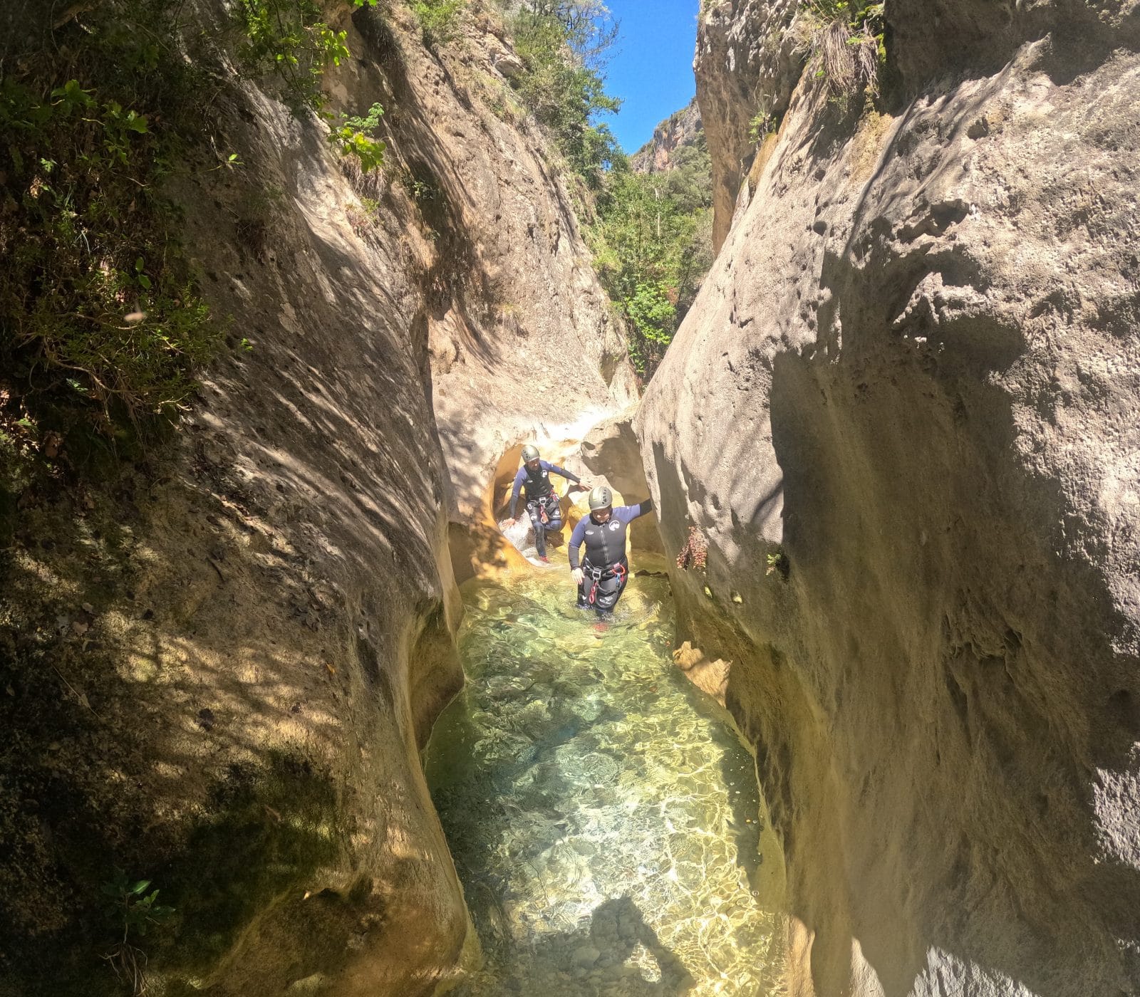 Two participants wading through a narrow crystal-clear pool between smooth rock walls in Barranco de Gurp