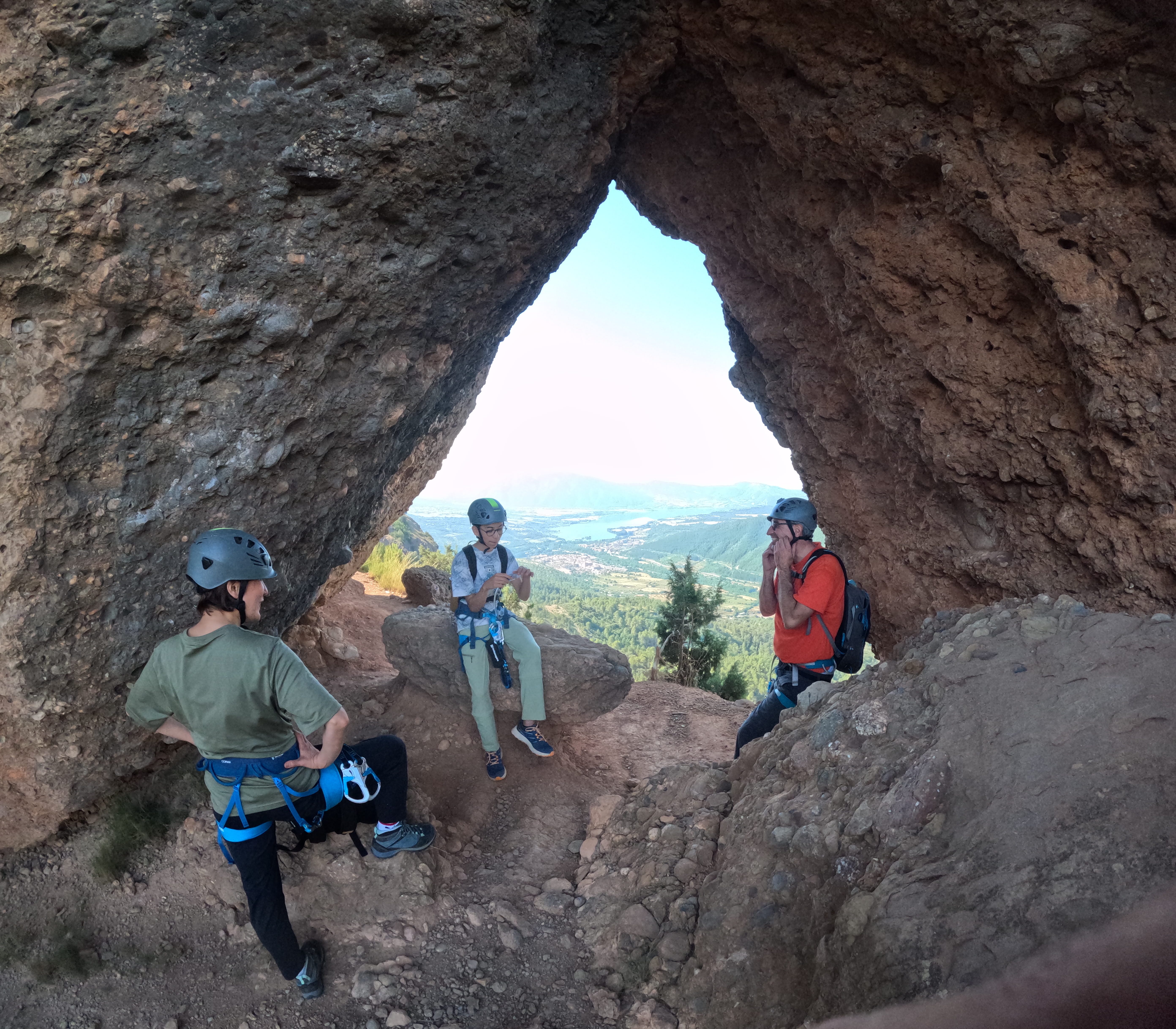 Participants taking a break inside the natural rock arch of the Vía Ferrata Roca Foradada, with panoramic views over the valley and reservoir near La Pobla de Segur