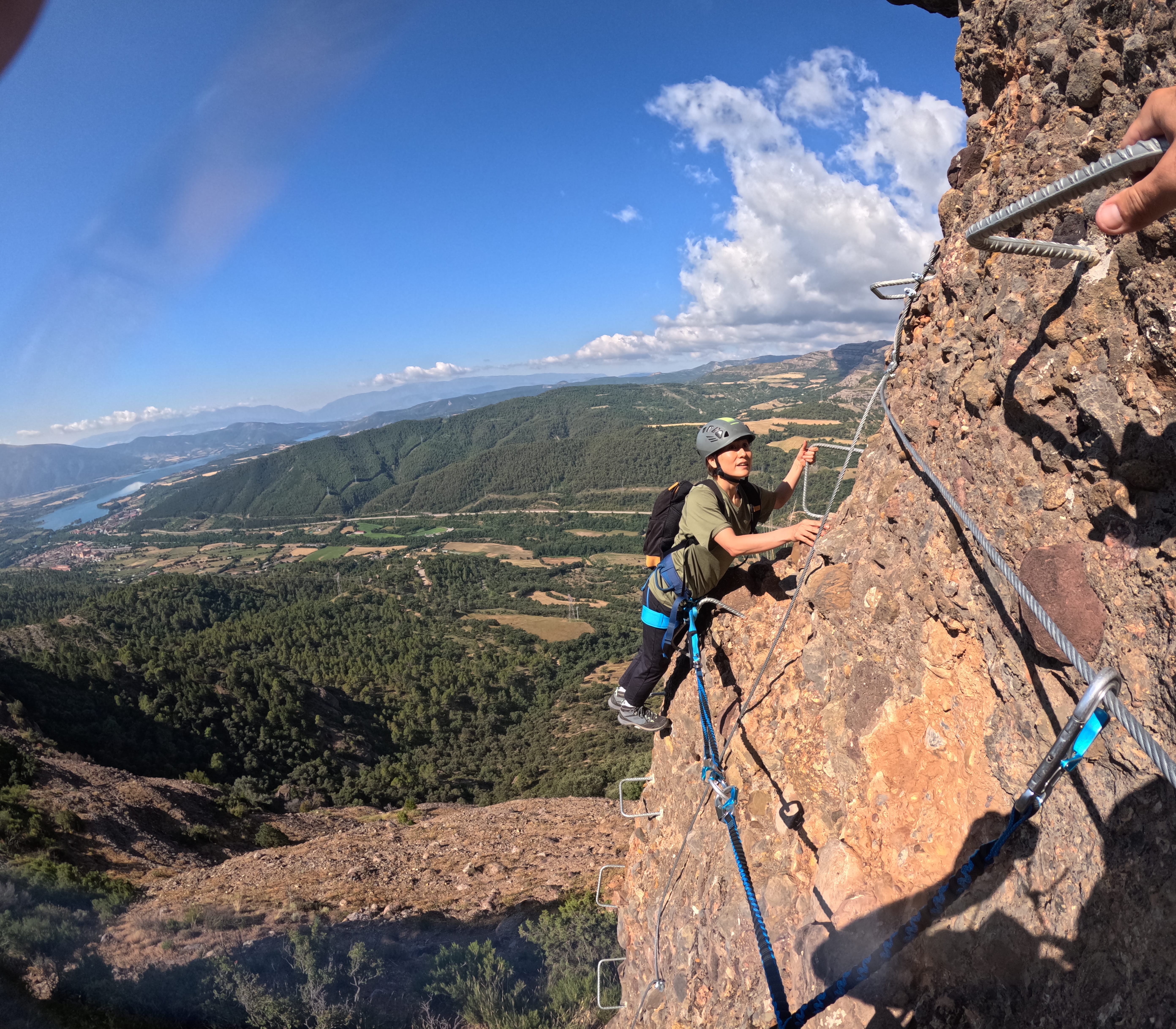 Climber ascending the exposed rock wall of the Vía Ferrata Roca Foradada using metal rungs and a safety cable, with panoramic views over La Pobla de Segur and the reservoir