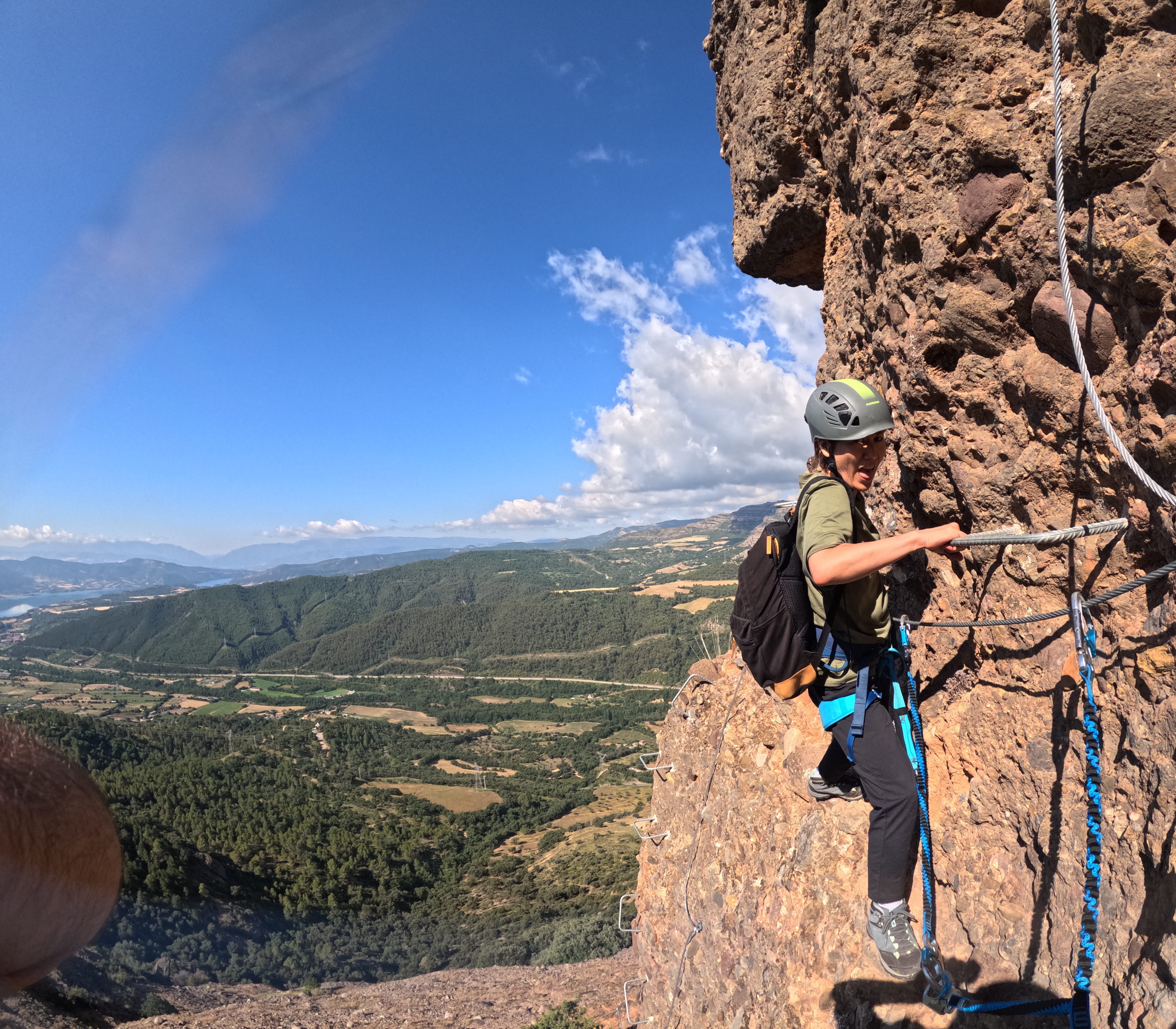 Climber traversing an exposed section of the Vía Ferrata Roca Foradada using metal rungs and a safety cable, with panoramic views over the valley near La Pobla de Segur