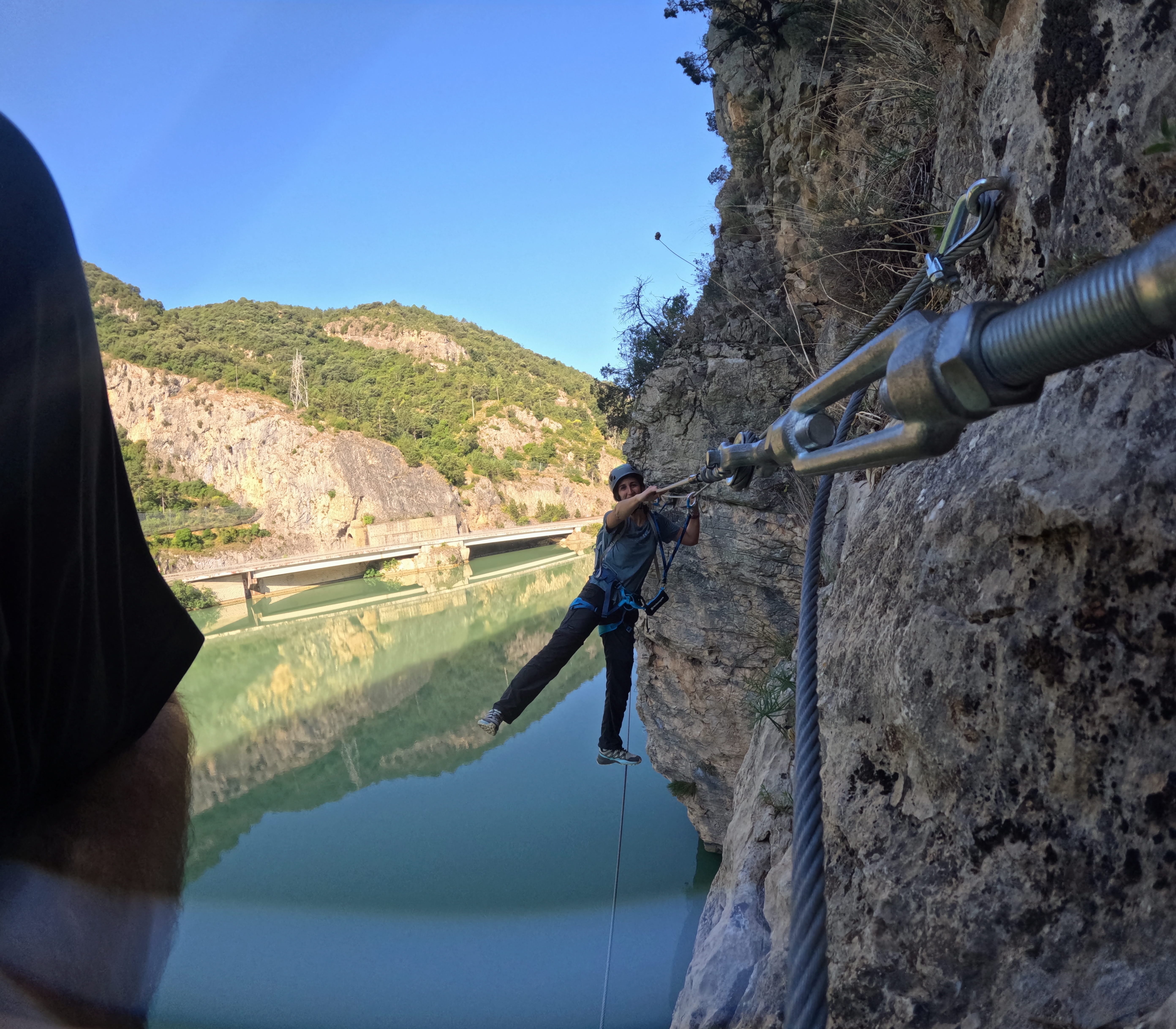 Participant crossing an exposed section of the vía ferrata Les Arcades, suspended above the reservoir and secured to the safety system with the rocky cliffs and mountains in the background