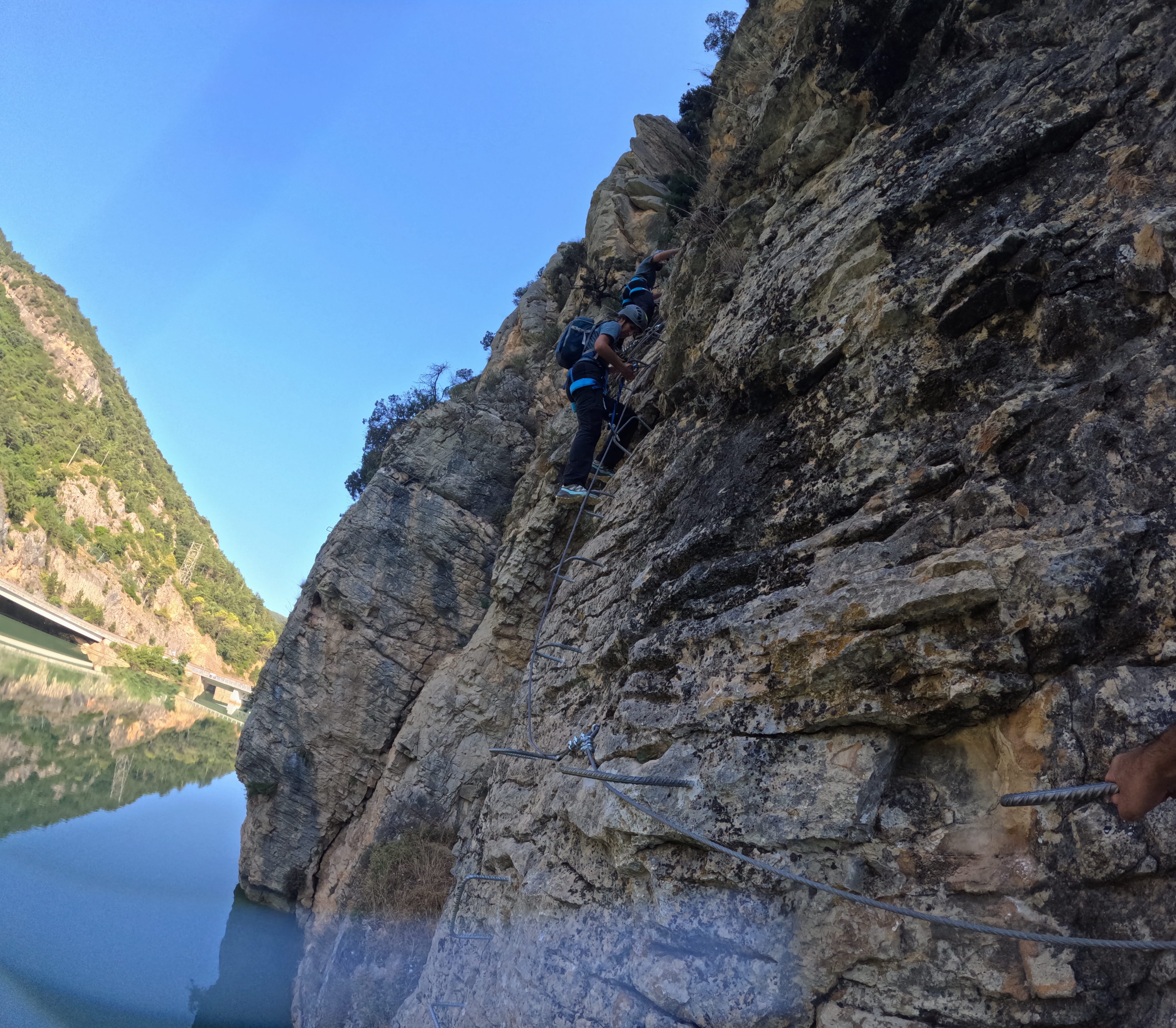 Participants climbing a steep section of the vía ferrata Les Arcades on metal rungs above the reservoir, secured to the safety cable along the rocky cliff