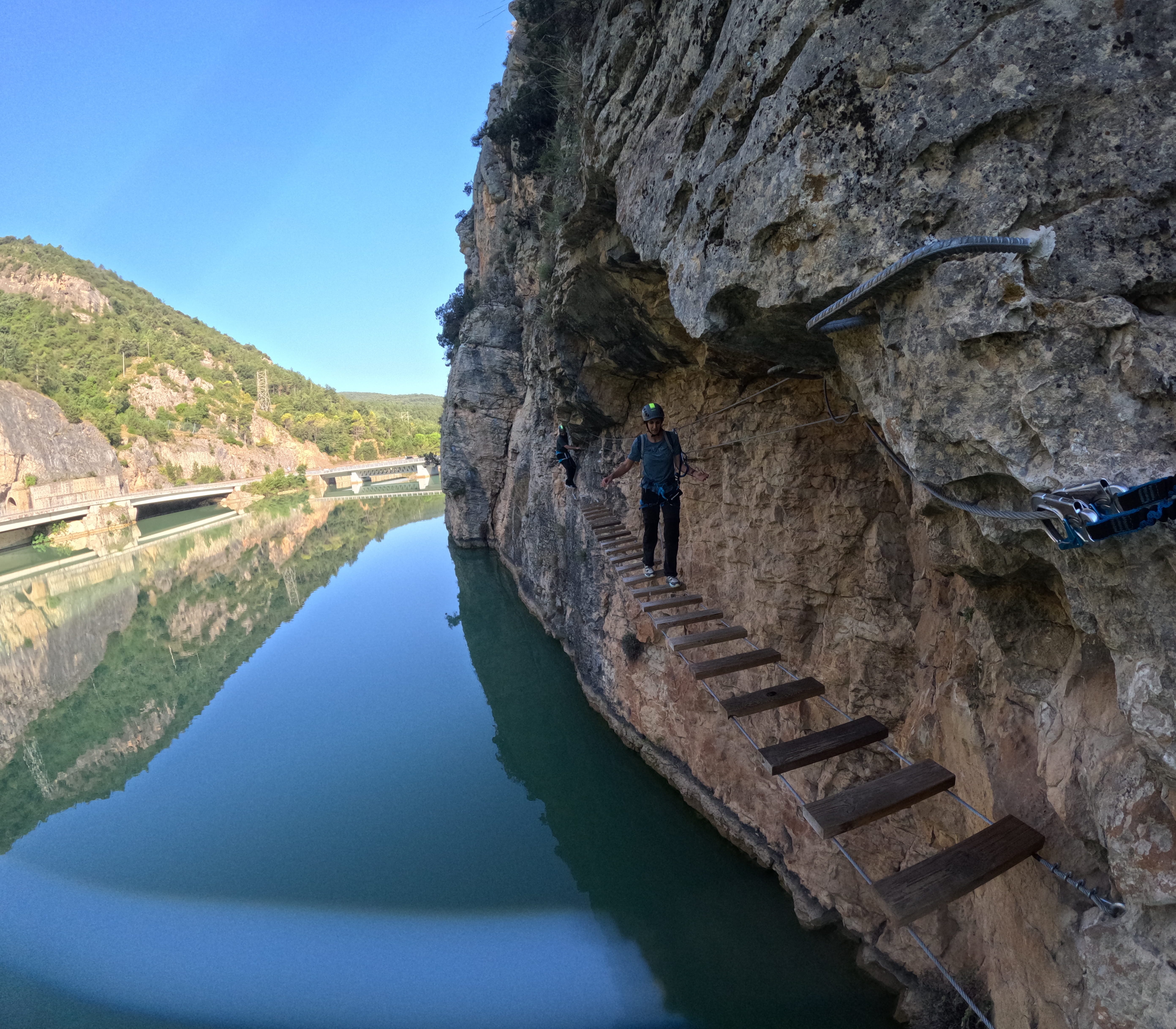 Participant crossing the suspended wooden bridge of the vía ferrata Les Arcades above the reservoir, secured to the safety cable with the rocky cliff and mountains in the background
