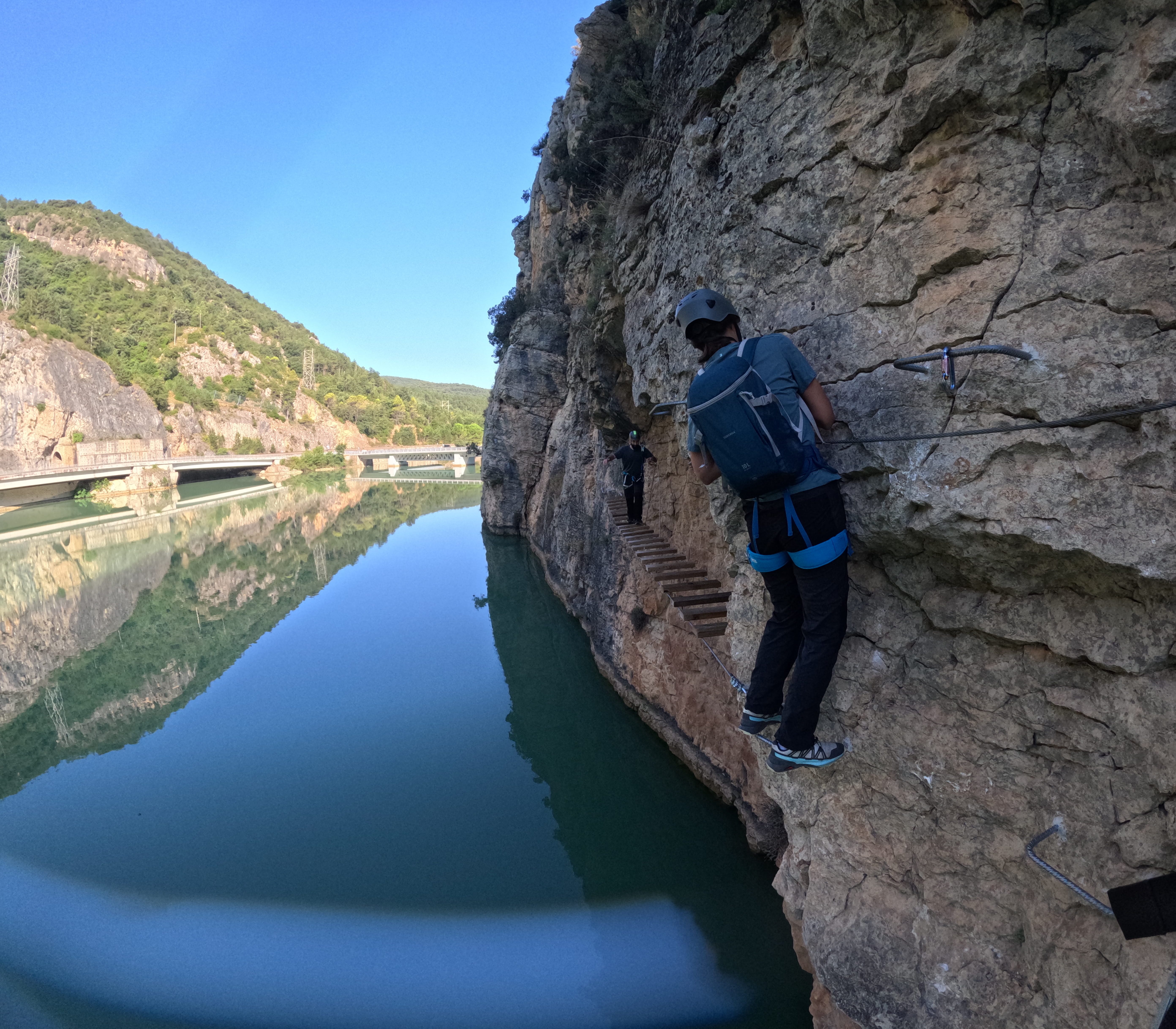 Participant traversing an exposed section of the vía ferrata Les Arcades above the reservoir, following the safety cable towards the suspended wooden bridge with mountain scenery in the background