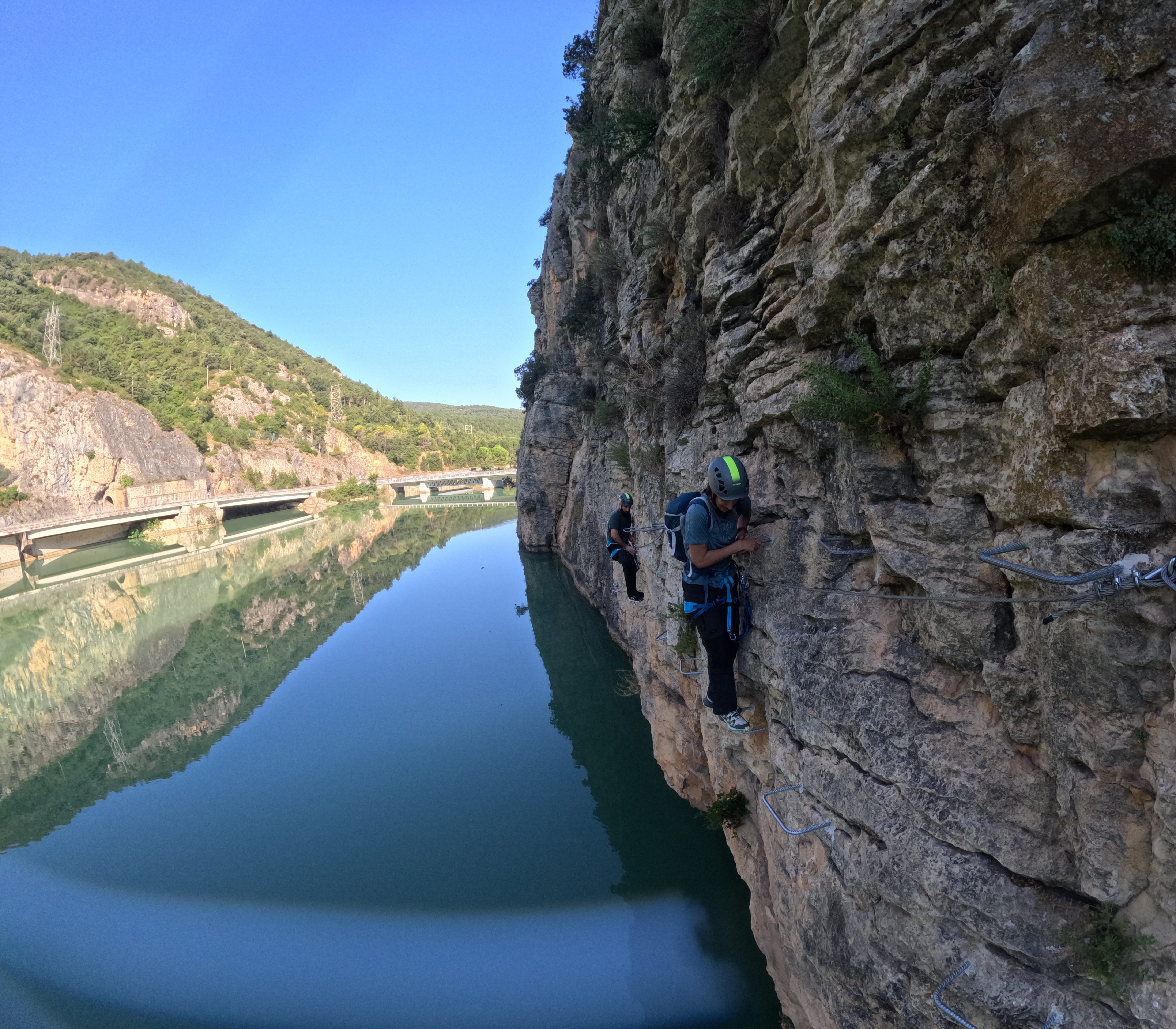 Participants traversing an exposed section of the vía ferrata Les Arcades on metal footholds along the rocky cliff above the reservoir, secured to the safety cable with mountains reflected in the water