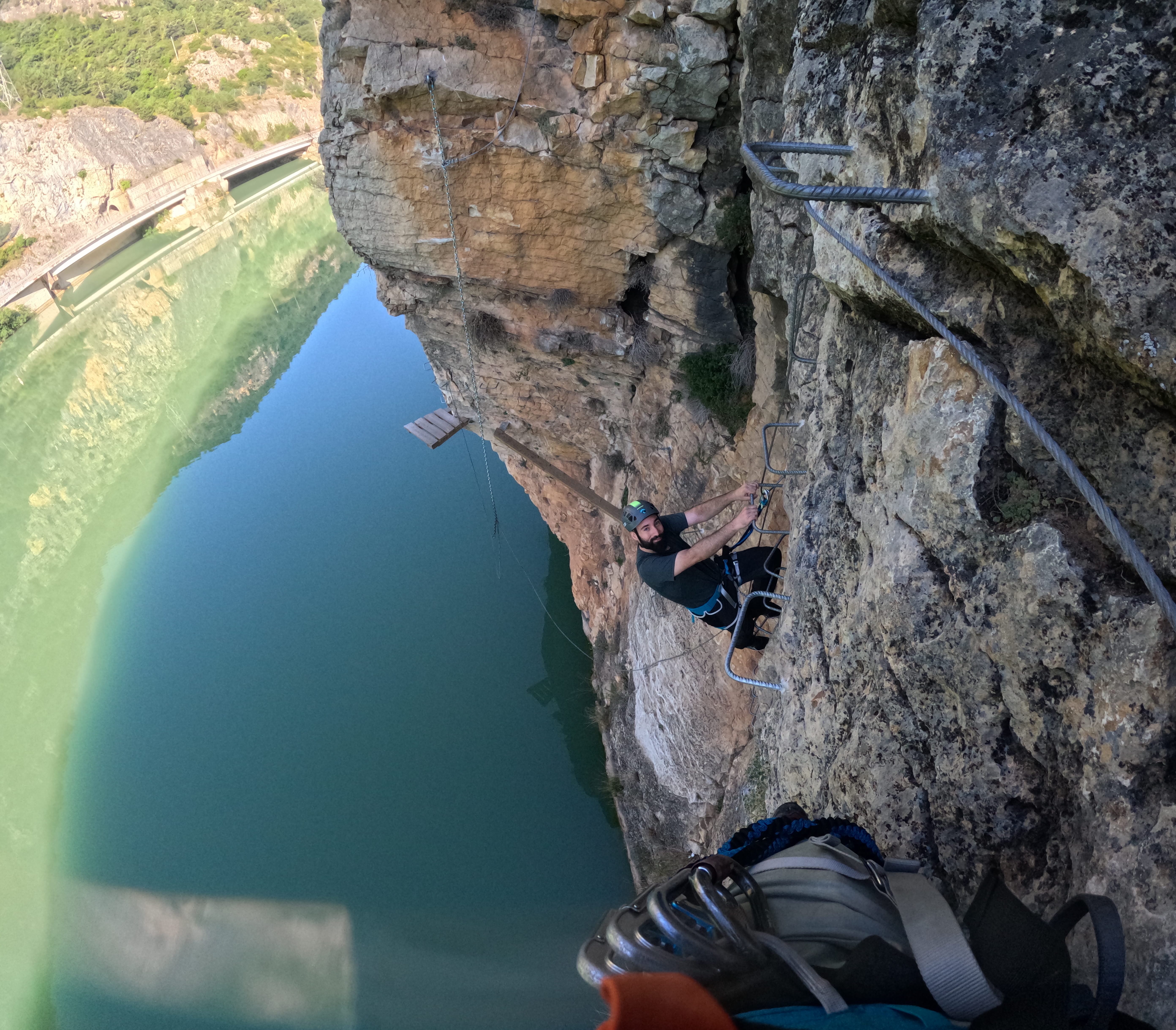 Participant climbing a vertical section of the vía ferrata Les Arcades on metal rungs above the reservoir, secured to the safety cable with the rocky cliff and water below