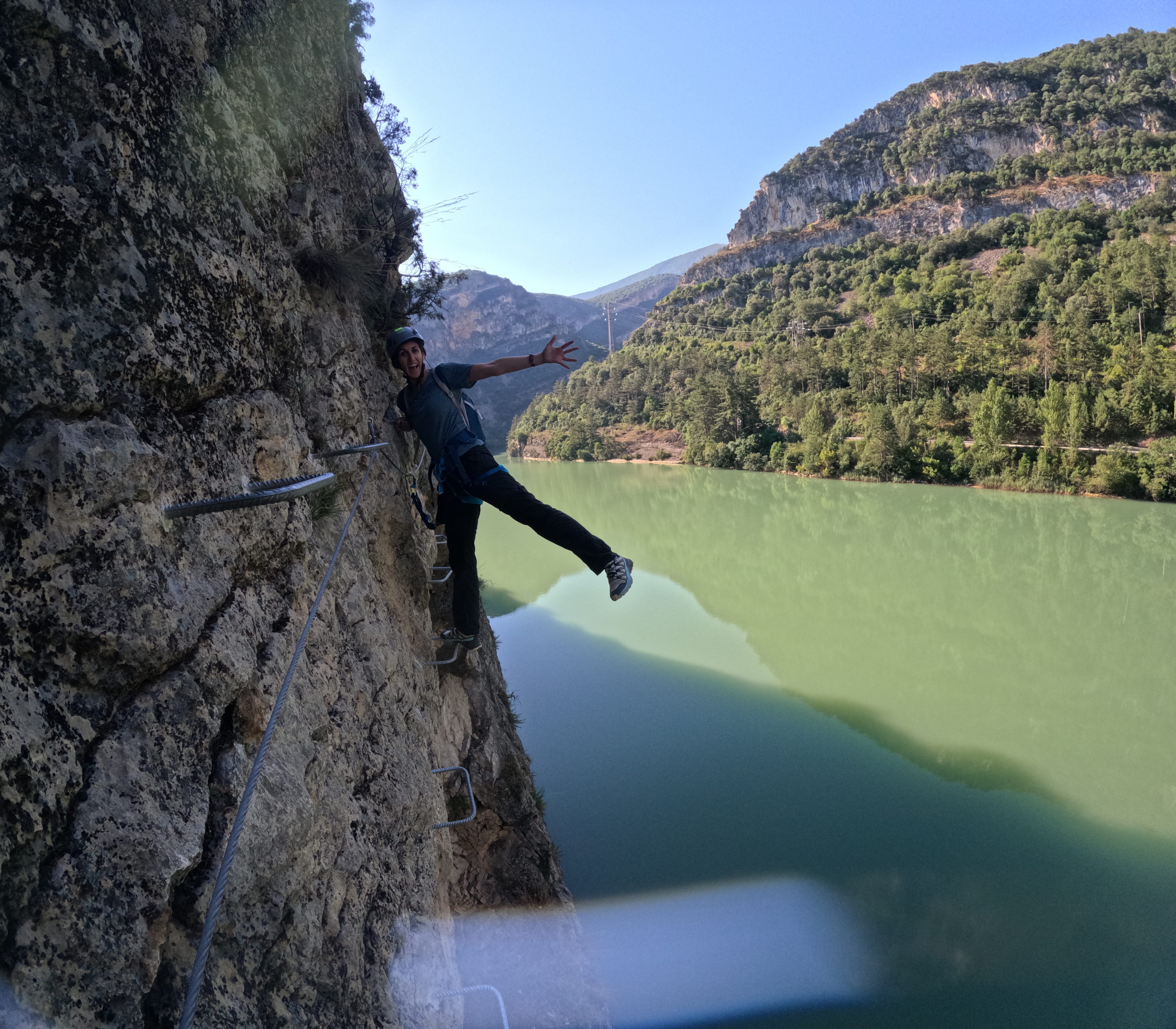 Participant enjoying an exposed section of the vía ferrata Les Arcades on metal footholds above the reservoir, secured to the safety cable with spectacular mountain scenery in the background