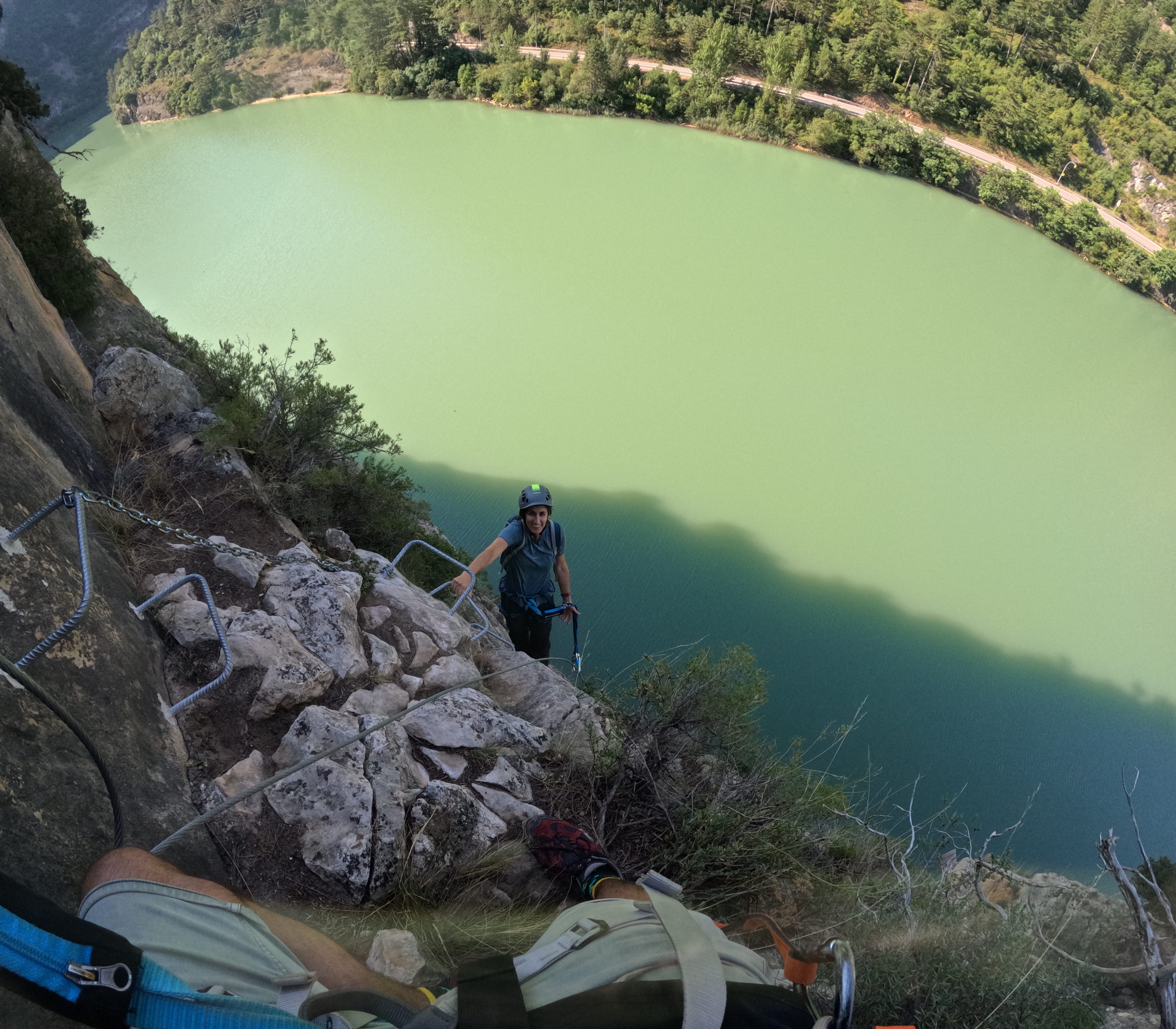 Participant climbing a steep section of the vía ferrata Les Arcades on metal rungs high above the reservoir, secured to the safety cable with an impressive view of the water and surrounding forest