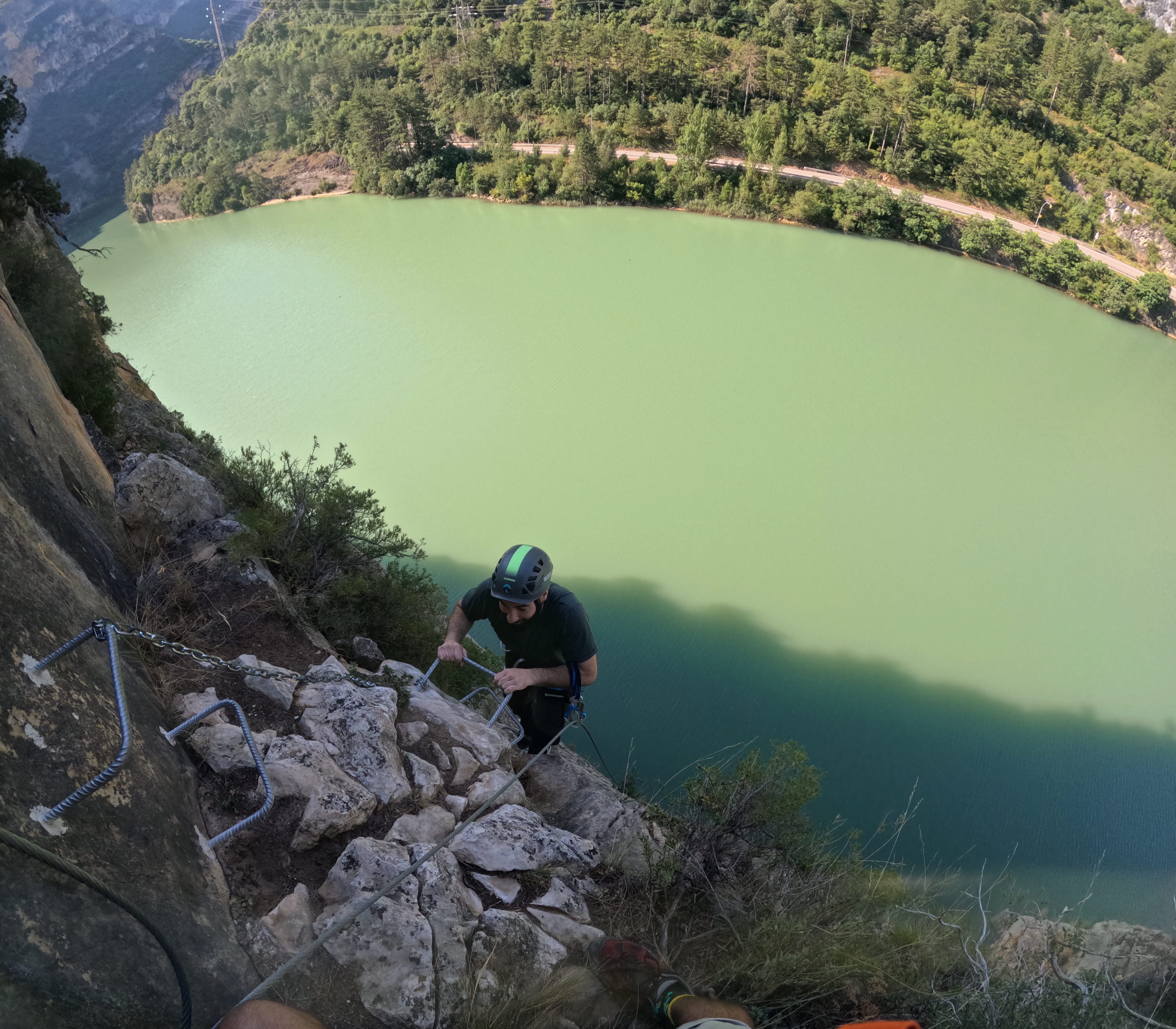 Participant climbing a steep section of the vía ferrata Les Arcades on metal rungs above the reservoir, secured to the safety cable with panoramic views of the water and surrounding forest