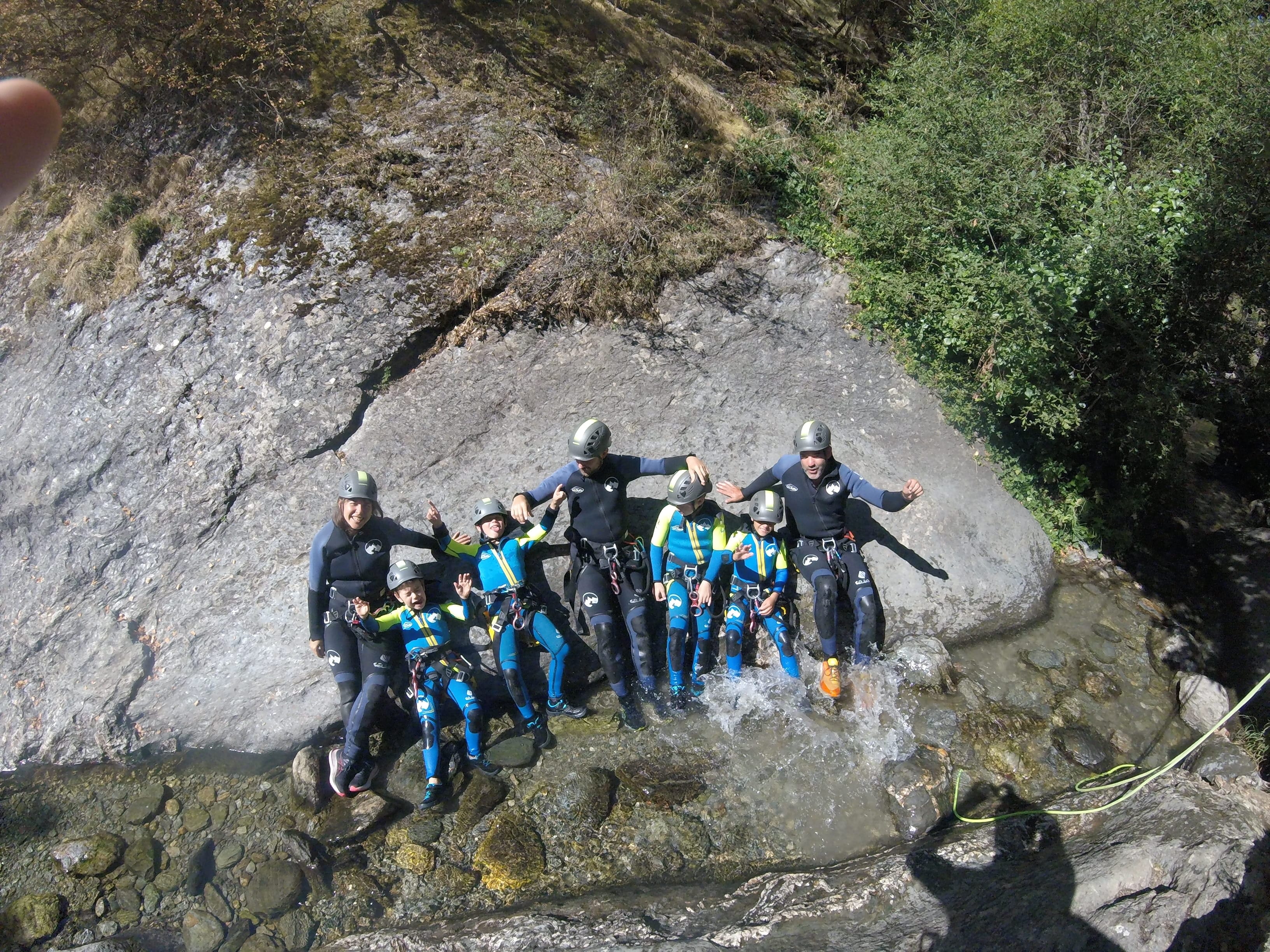 Group of adults and children posing together in canyoning gear beside a rocky stream during a family descent of Barranco de Berrós