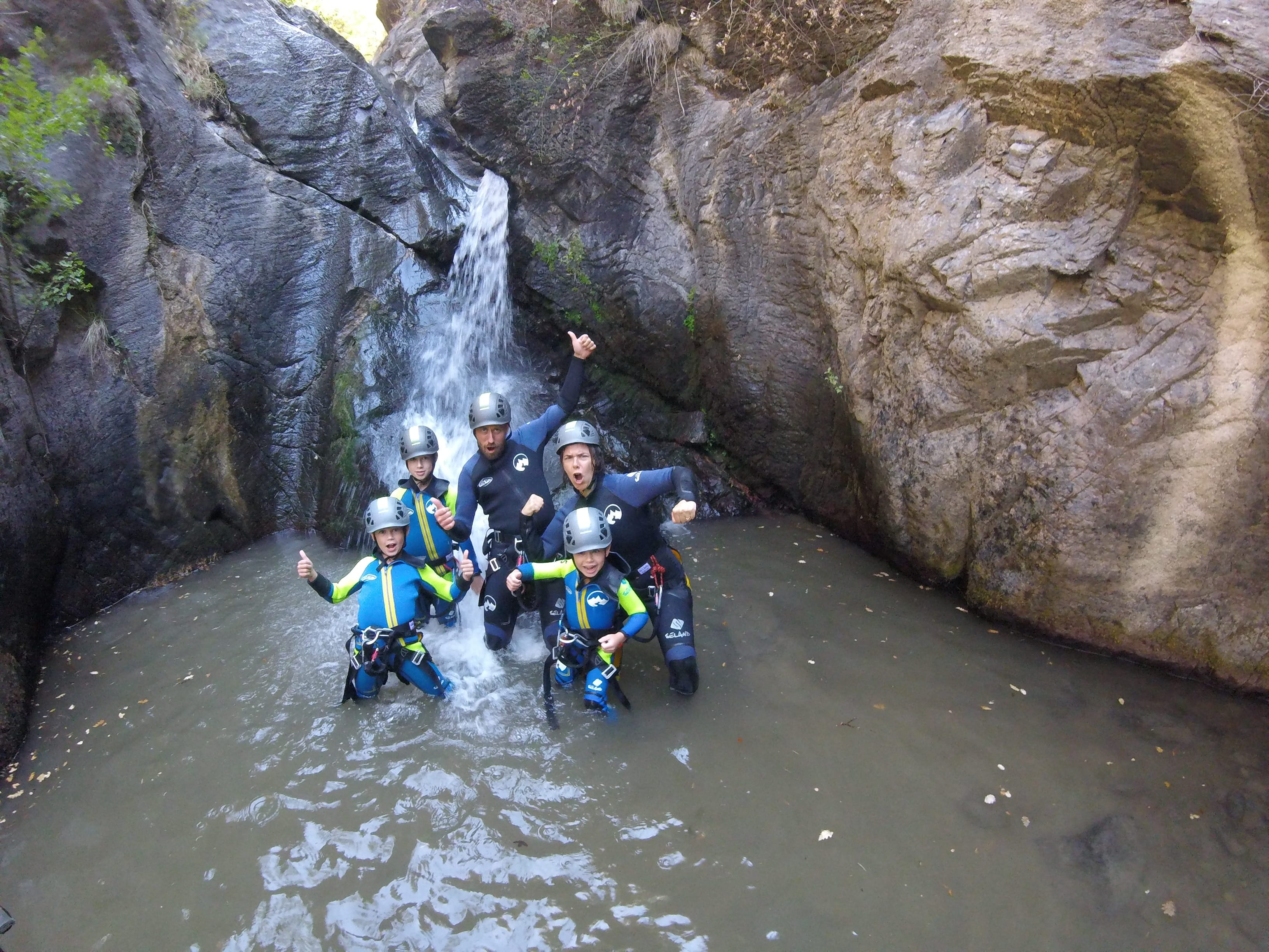 Adults and children posing in a natural pool beneath a waterfall during a family canyoning descent in Barranco de Berrós