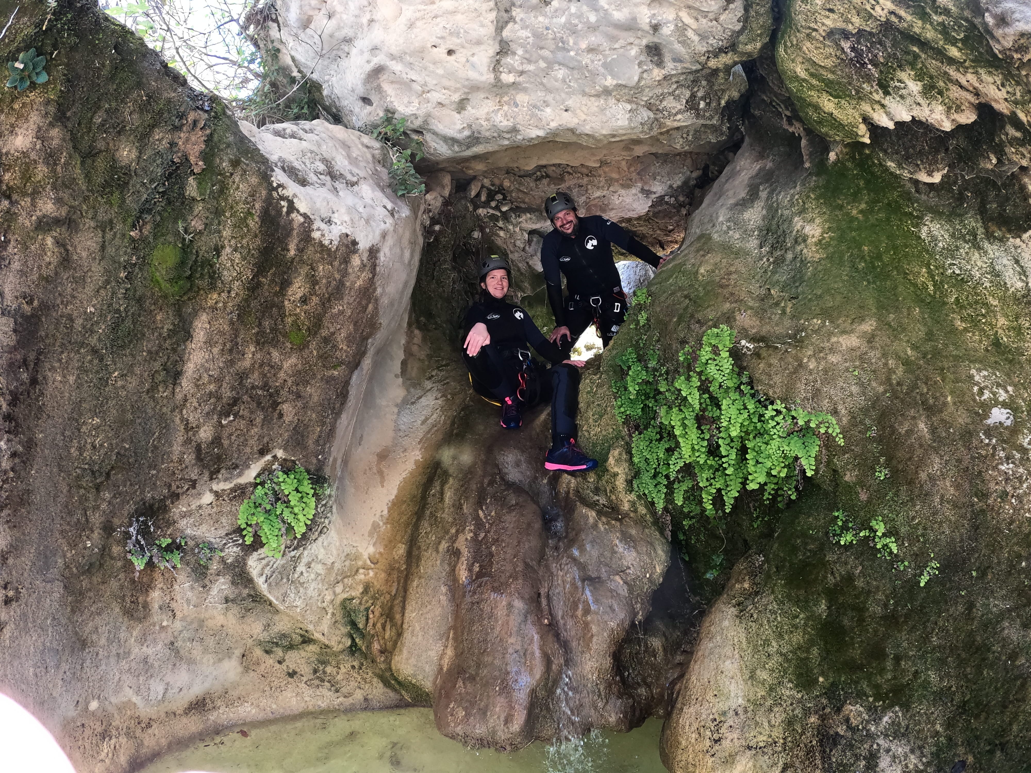 Two participants descending through a narrow moss-covered rocky passage with a small pool in Barranco de Gurp