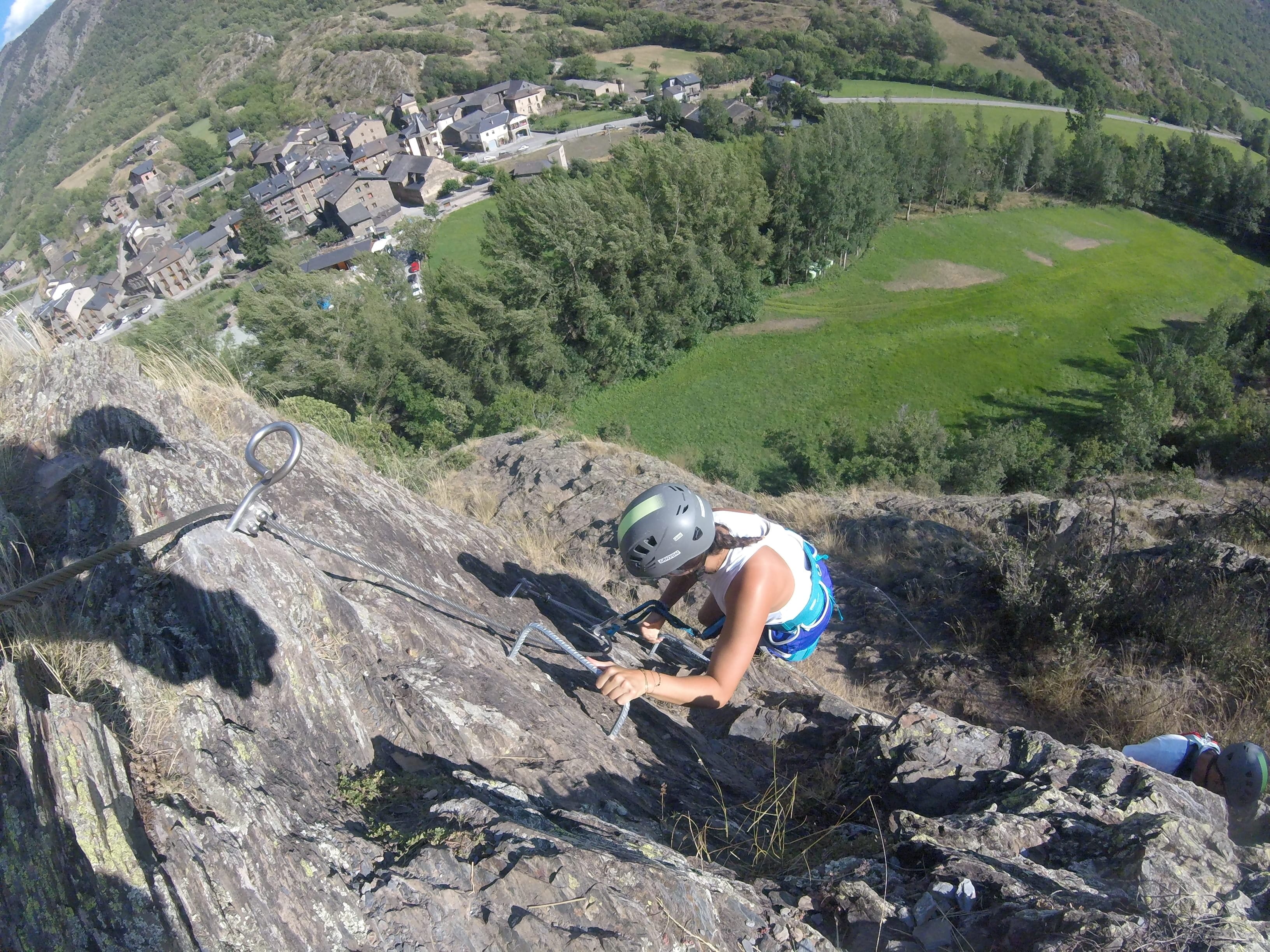 Participante subiendo por un tramo rocoso y empinado de la vía ferrata Salt de Botanal, con cable de seguridad y vistas al pueblo y al valle verde al fondo