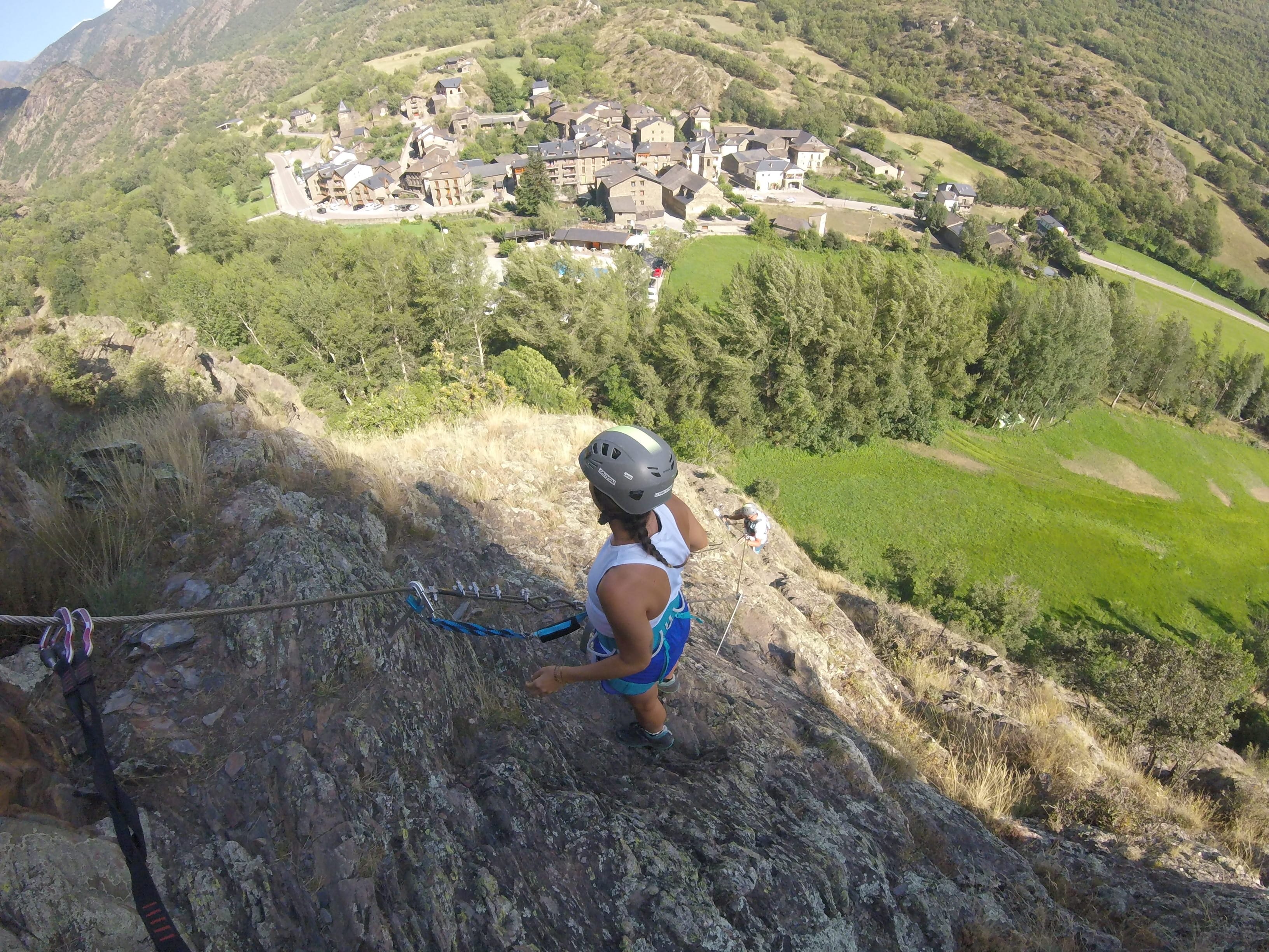 Participante en un tramo rocoso y empinado de la vía ferrata Salt de Botanal, asegurada al cable de seguridad con vistas panorámicas al pueblo de montaña y al valle verde al fondo