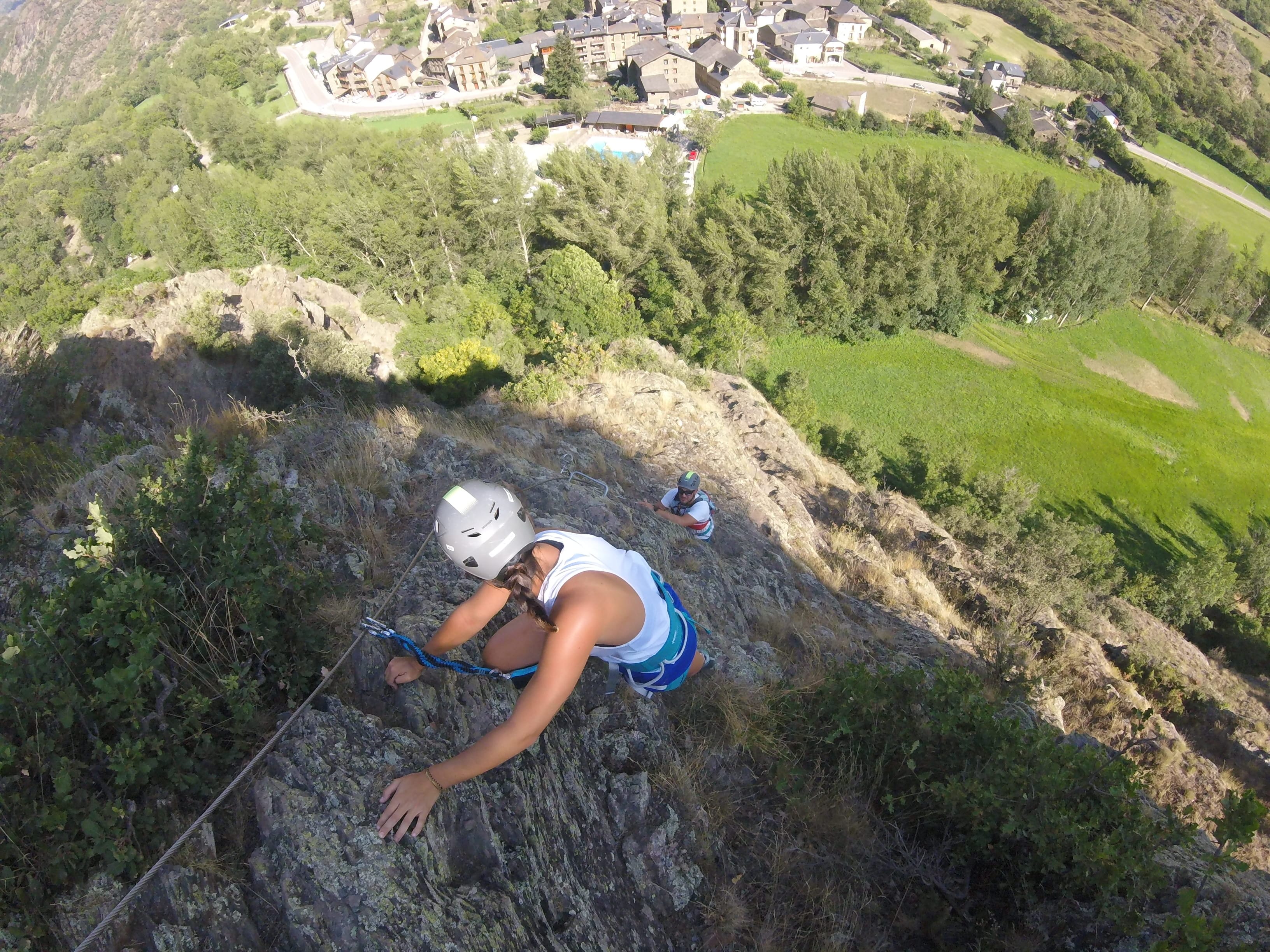 Participante subiendo por un tramo rocoso y empinado de la vía ferrata Salt de Botanal, asegurada al cable de seguridad con vistas al pueblo y al valle verde al fondo