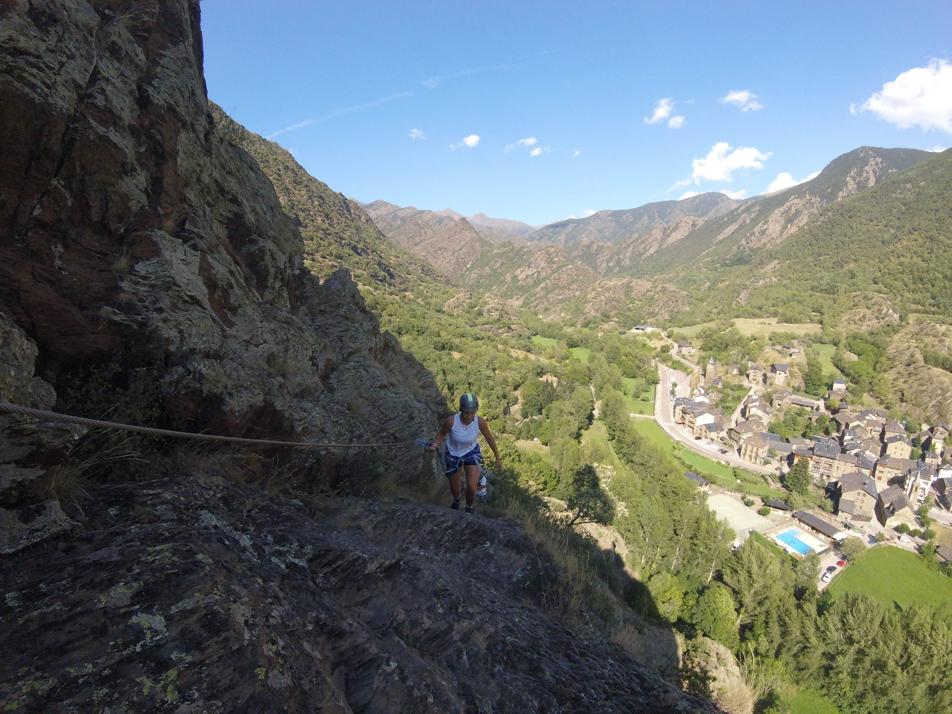 Participante subiendo por la vía ferrata Salt de Botanal en una ladera rocosa, con vistas panorámicas al pueblo y al valle pirenaico
