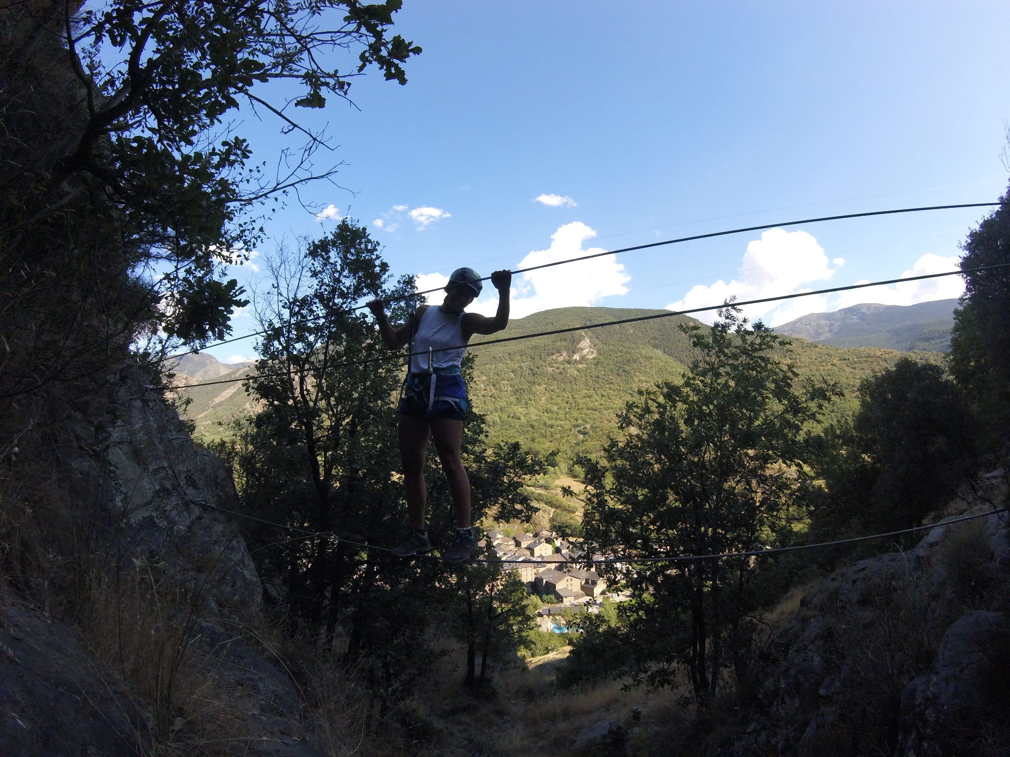 Participante cruzando un puente de cables en la vía ferrata Salt de Botanal, con paisaje de montaña y el pueblo visible en el valle al fondo