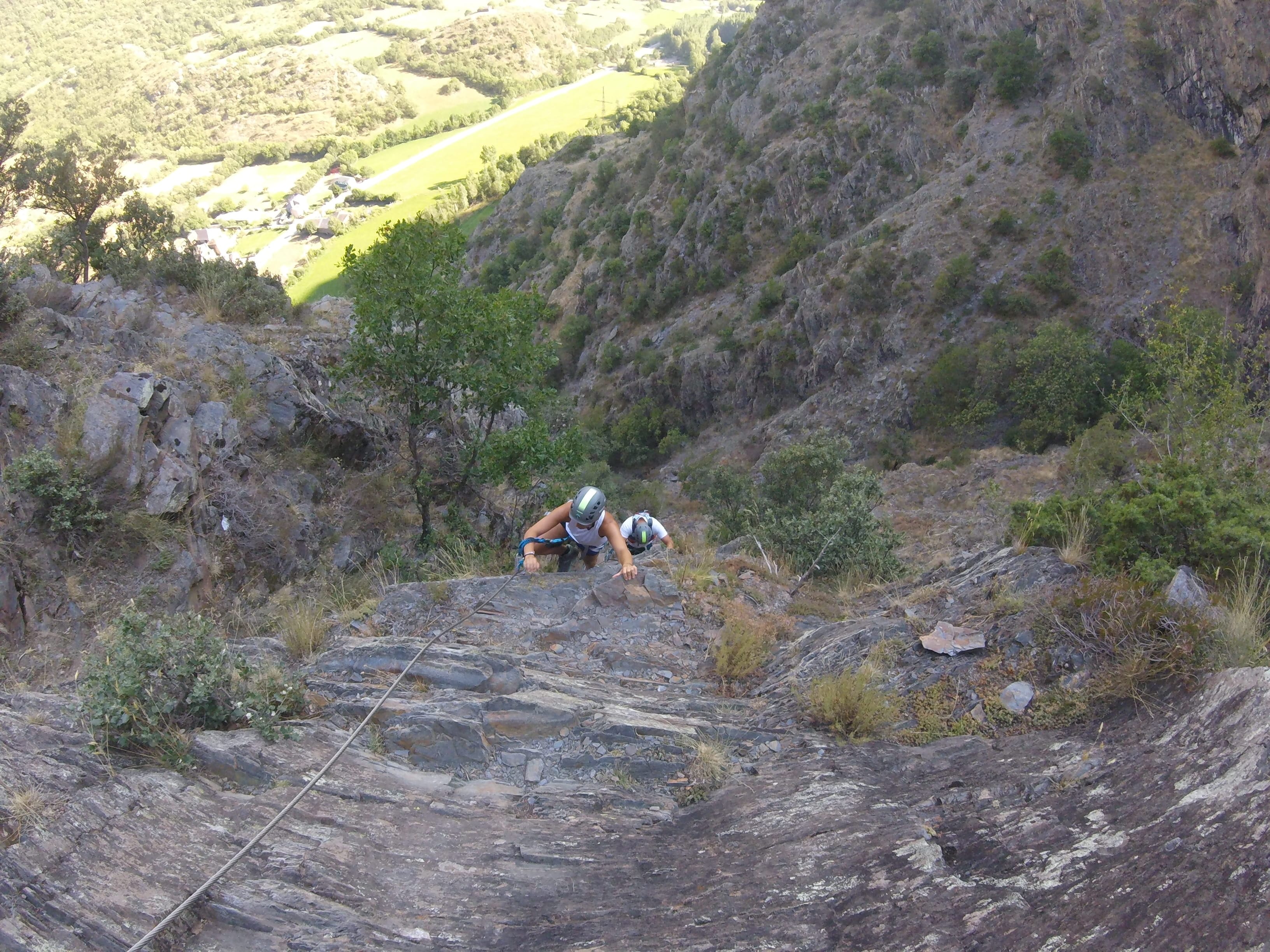 Participantes subiendo por un tramo rocoso y empinado de la vía ferrata Salt de Botanal, asegurados al cable de seguridad con el valle de montaña al fondo