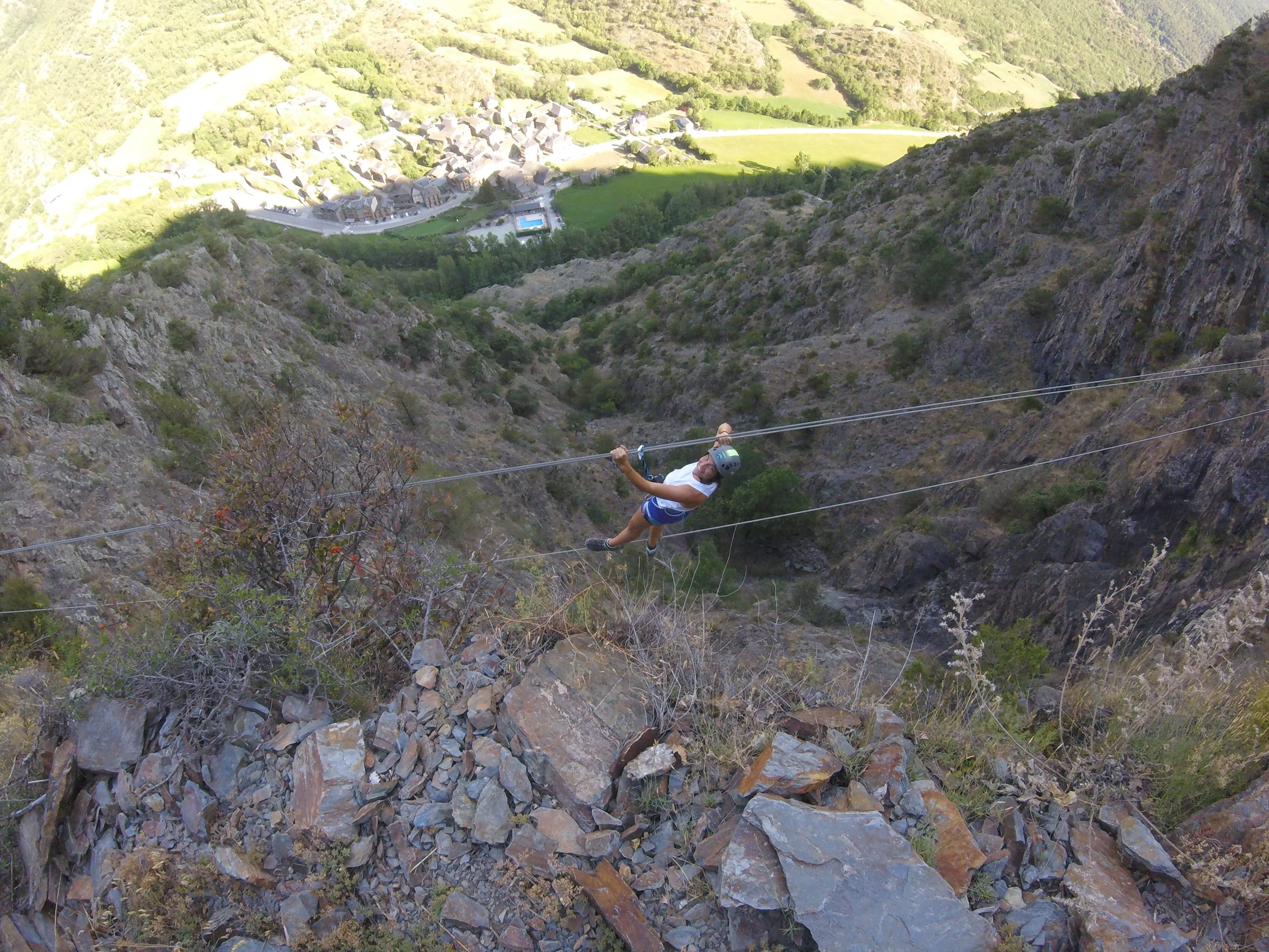 Participante cruzando un puente de cables elevado en la vía ferrata Salt de Botanal, suspendida sobre un barranco rocoso con vistas panorámicas al pueblo y al valle al fondo
