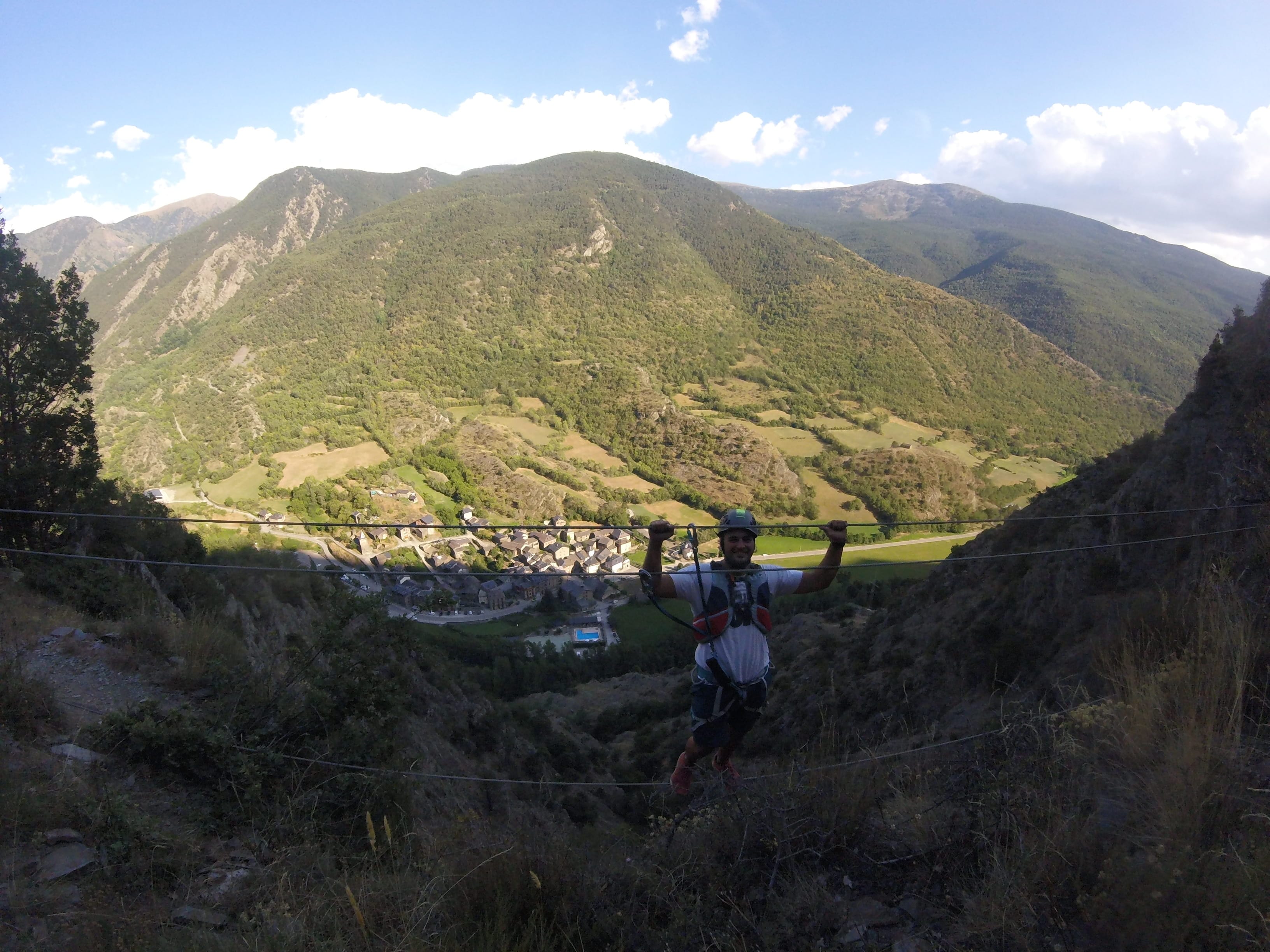 Participante cruzando un puente de cables elevado en la vía ferrata Salt de Botanal, con vistas panorámicas a las montañas pirenaicas, el valle y el pueblo al fondo