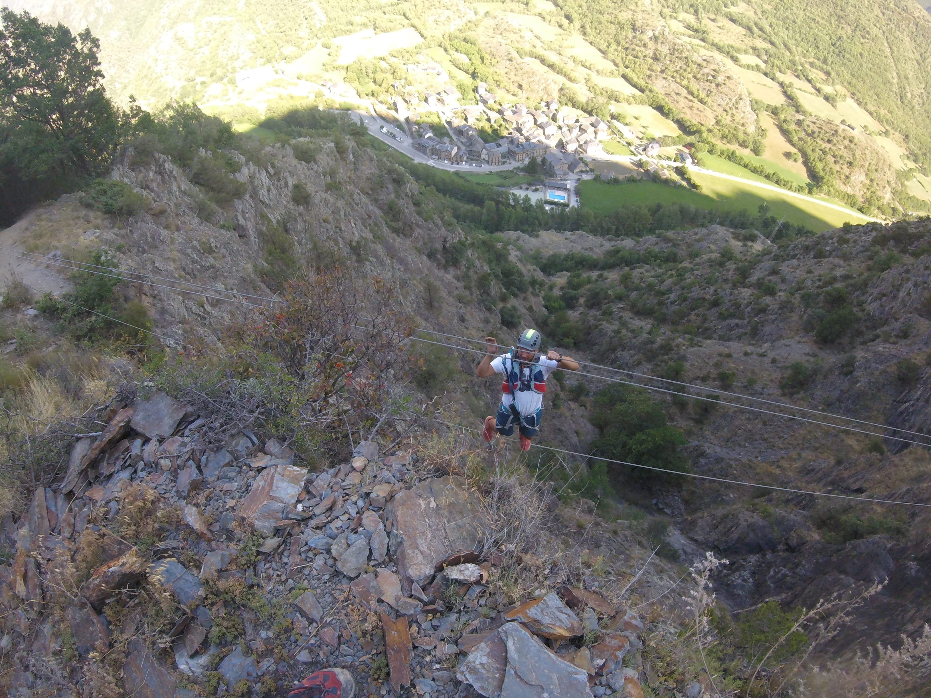 Participante cruzando un puente de cables elevado en la vía ferrata Salt de Botanal, suspendido sobre un barranco rocoso con el pueblo de montaña visible al fondo