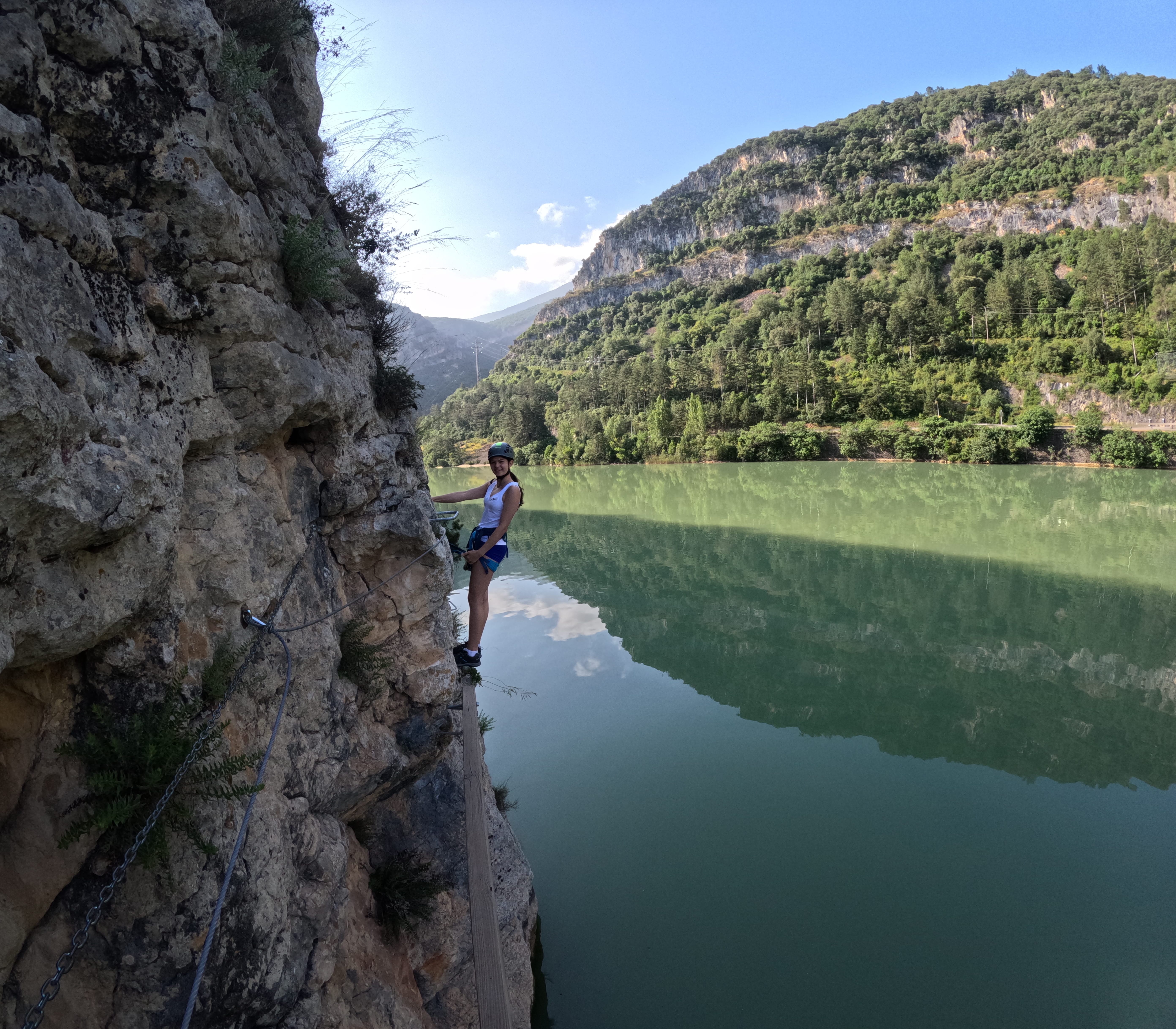 Participant traversing an exposed section of the vía ferrata Les Arcades along the rocky cliff above the reservoir, secured to the safety cable, with forested mountains reflected in the calm water