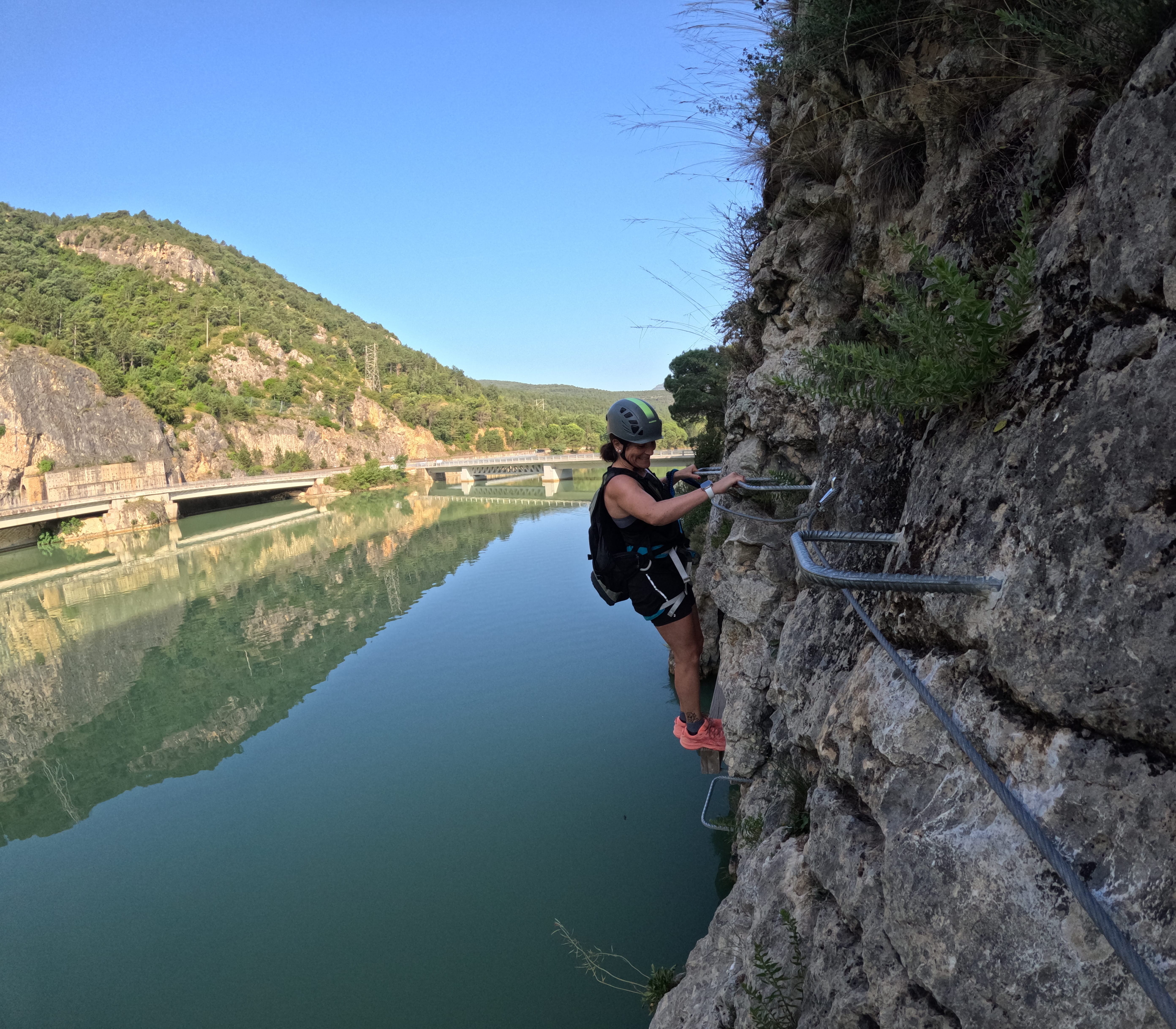 Participant traversing an exposed section of the vía ferrata Les Arcades on metal footholds above the reservoir, secured to the safety cable, with mountains and a bridge reflected in the calm water