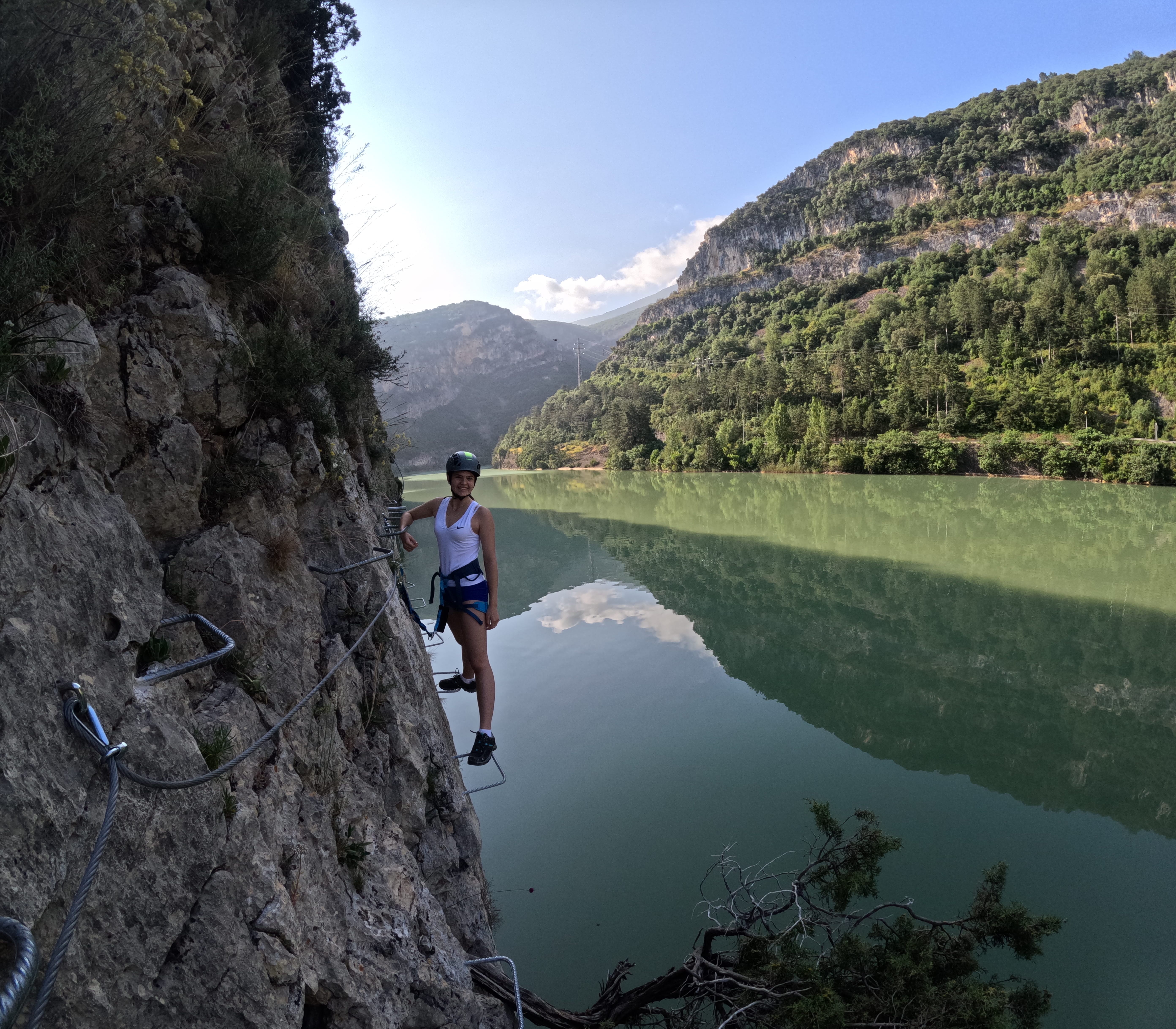 Participant traversing an exposed section of the vía ferrata Les Arcades on metal footholds above the reservoir, secured to the safety cable, with panoramic views of the forested mountains reflected in the water