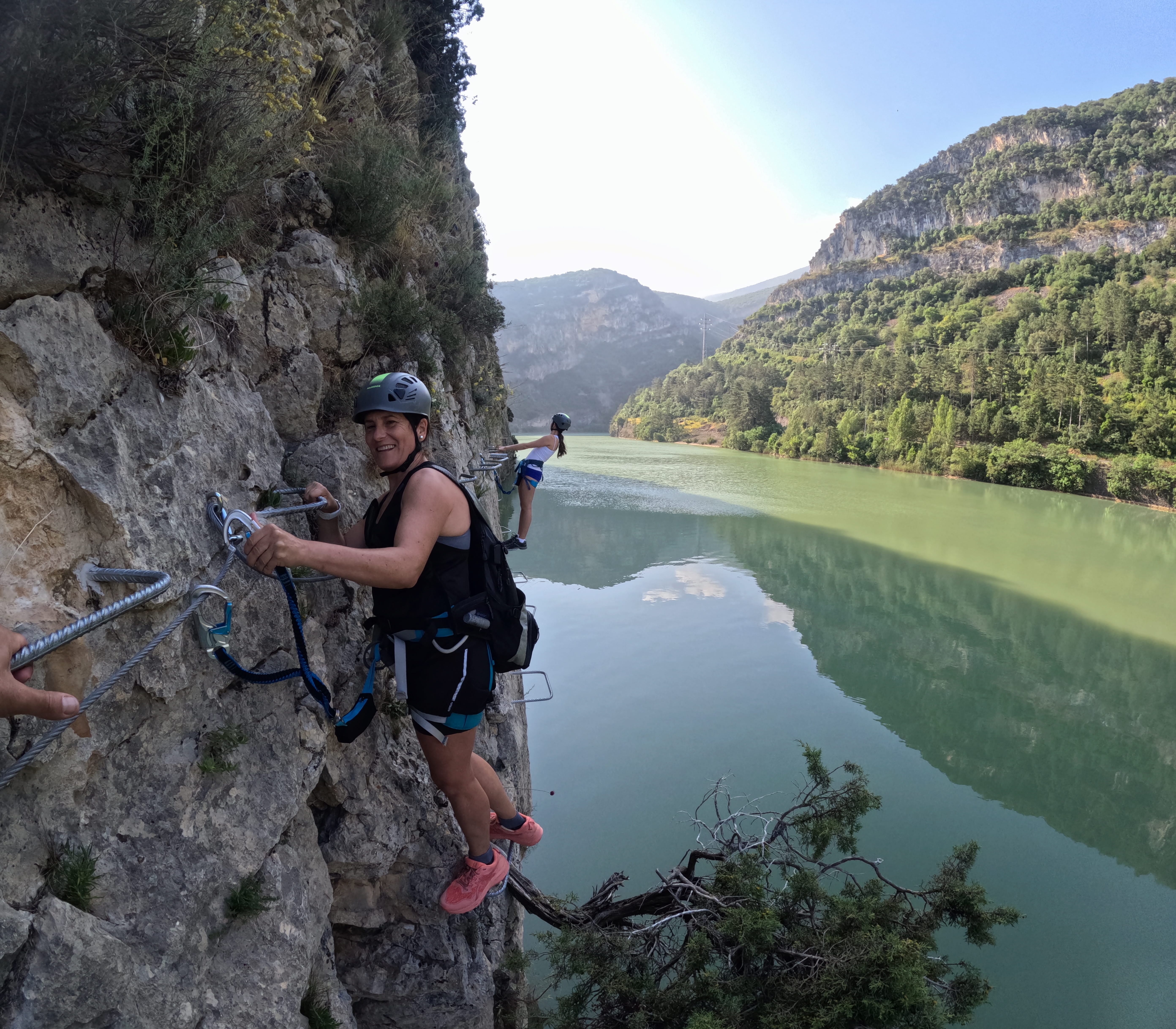 Participant on the vía ferrata Les Arcades progressing along metal footholds above the reservoir, secured to the safety cable, with another participant ahead and a panoramic mountain landscape in the background
