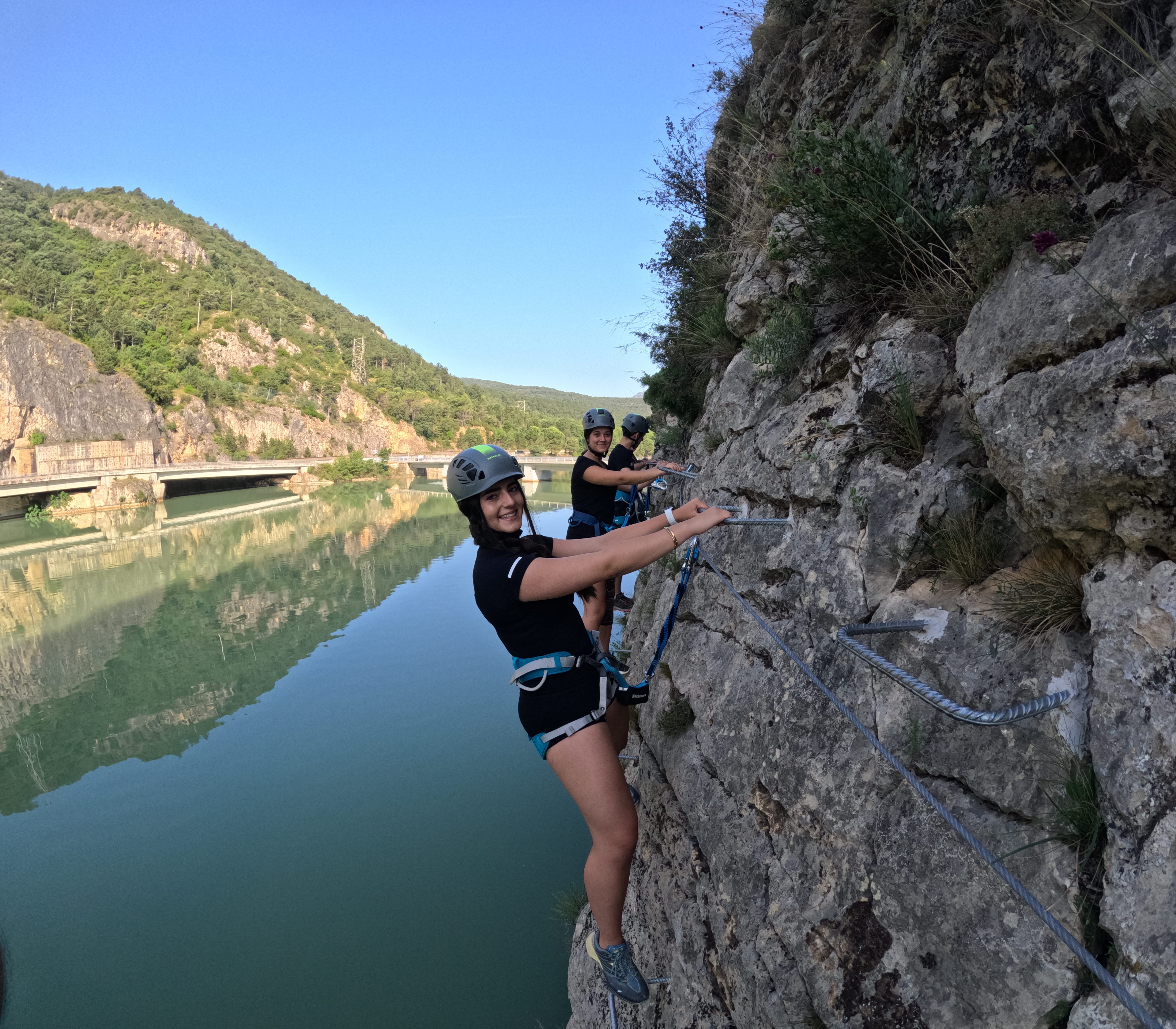 Participants on the vía ferrata Les Arcades traversing a rocky wall above the reservoir, secured to the safety cable, with mountains and reflections on the water in the background