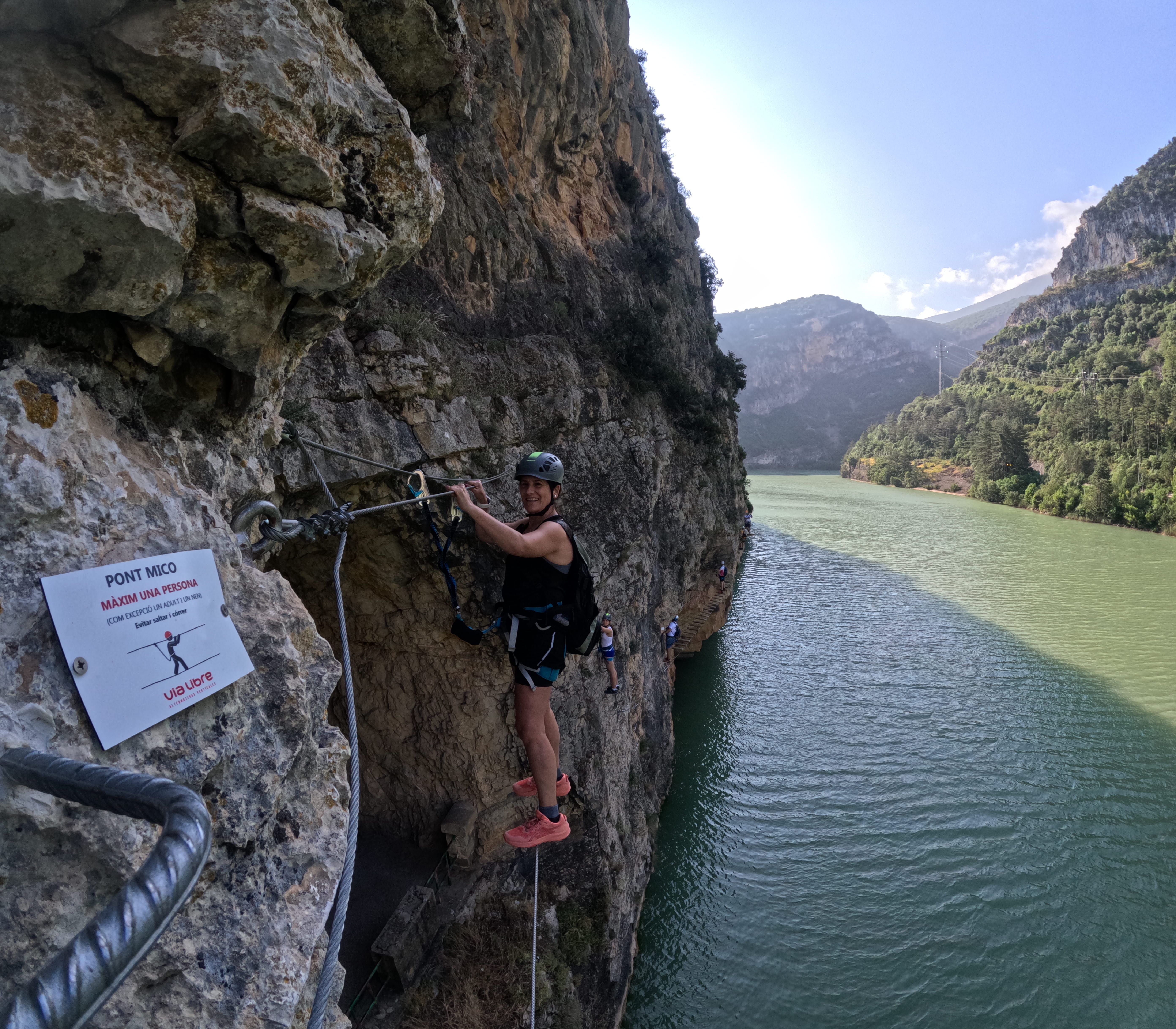 Participant crossing a short rope bridge on the vía ferrata Les Arcades above the reservoir, secured to the safety cable, with the rocky gorge and mountain landscape in the background