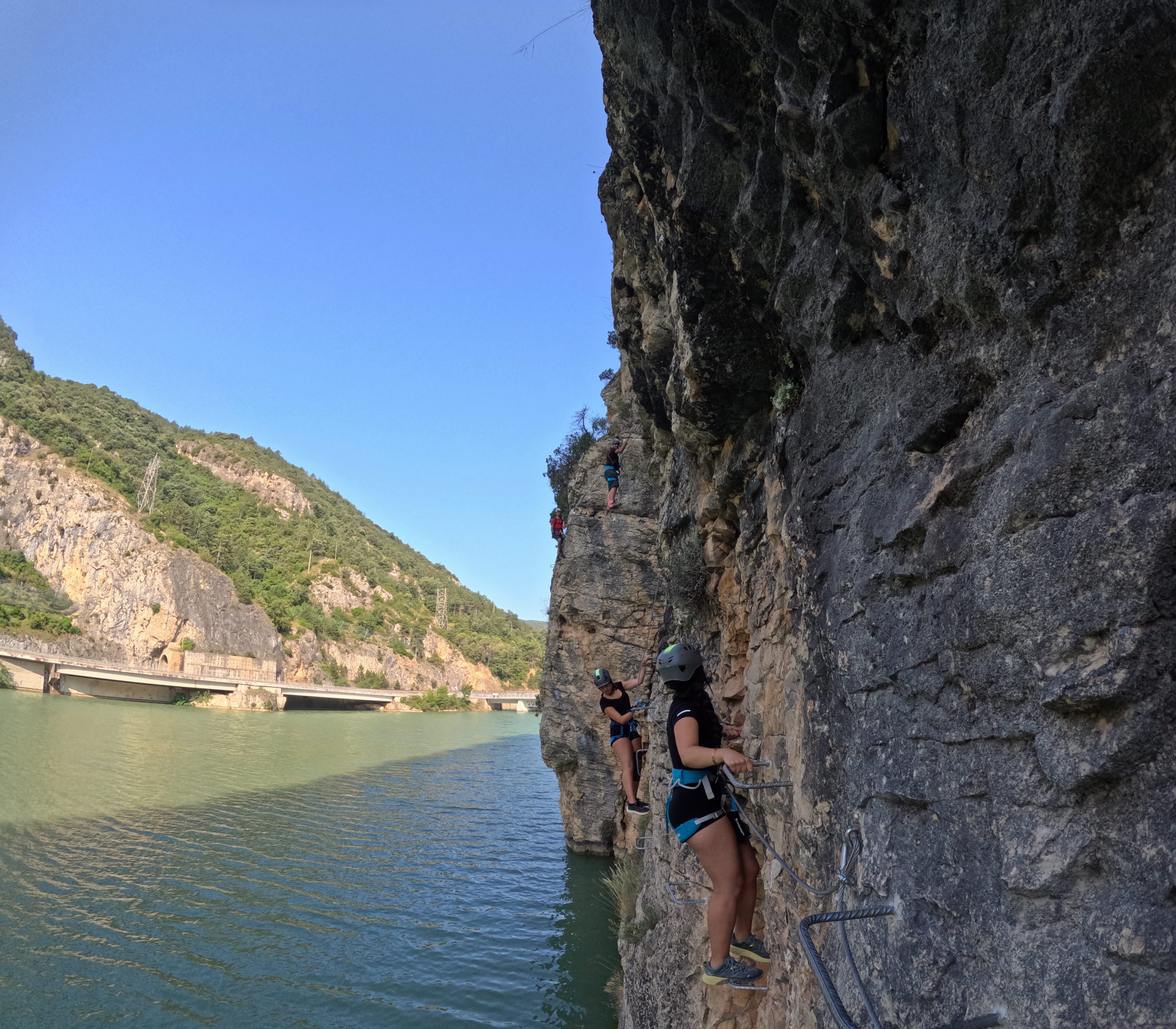Participants traversing the rocky wall on the vía ferrata Les Arcades above the reservoir, secured to the safety cable and using metal footholds, with mountains and a bridge in the background
