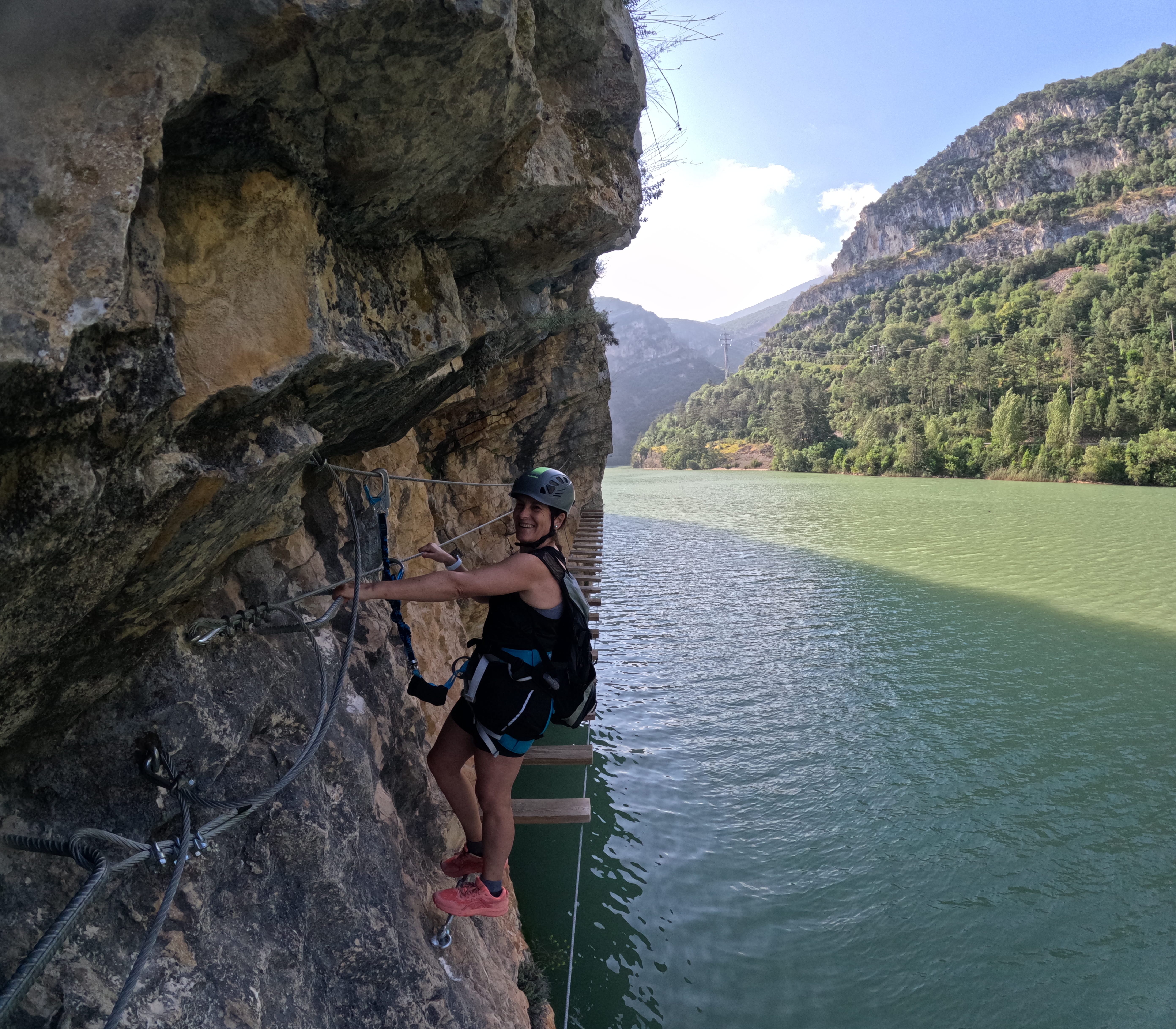 Participant on the vía ferrata Les Arcades traversing a rocky section above the reservoir, secured to the safety cable, with a suspended wooden walkway and mountain landscape in the background