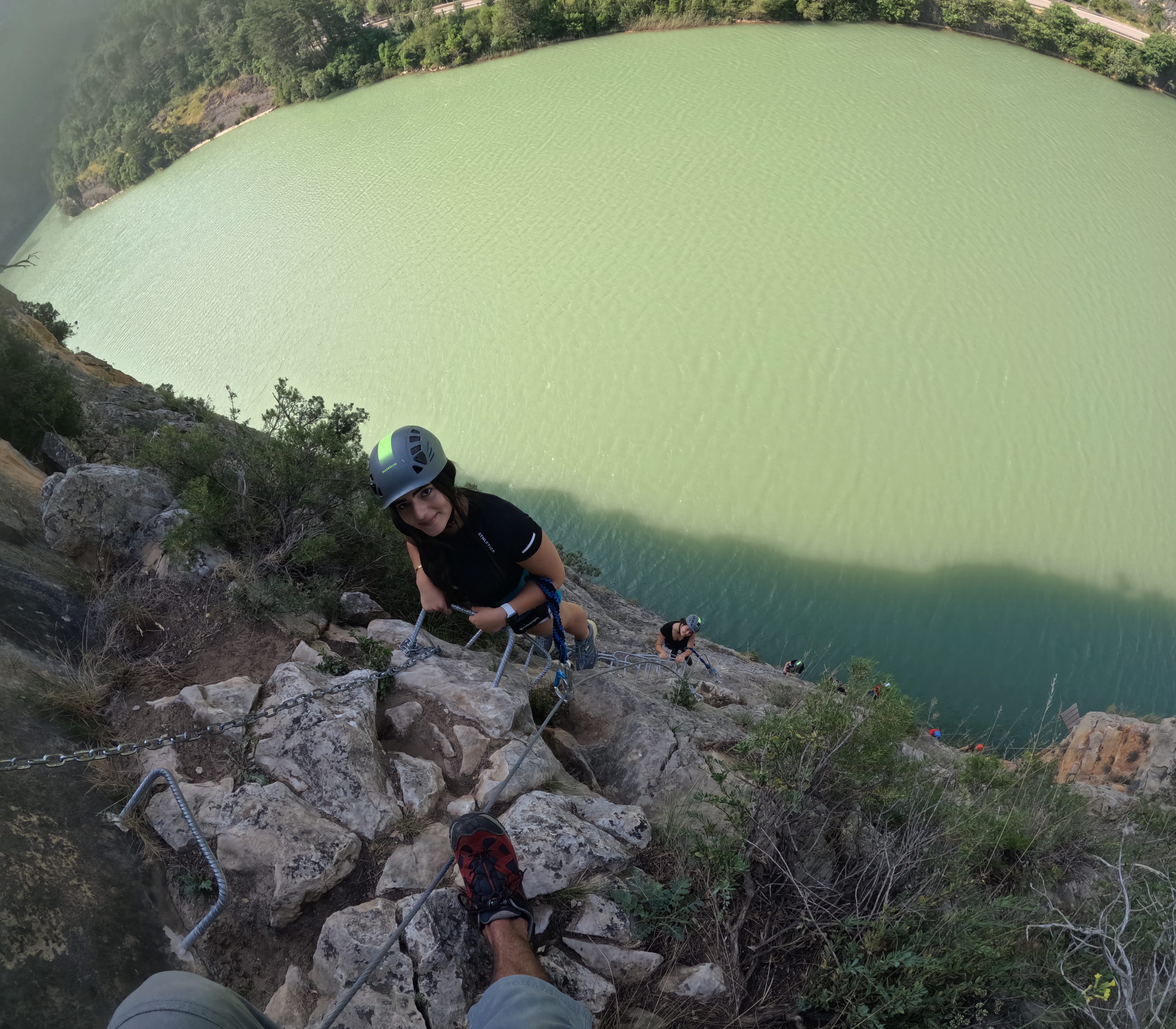 Participant climbing a steep section of the vía ferrata Les Arcades above the reservoir, secured to the safety cable and using metal footholds, with other participants visible below and panoramic views over the water
