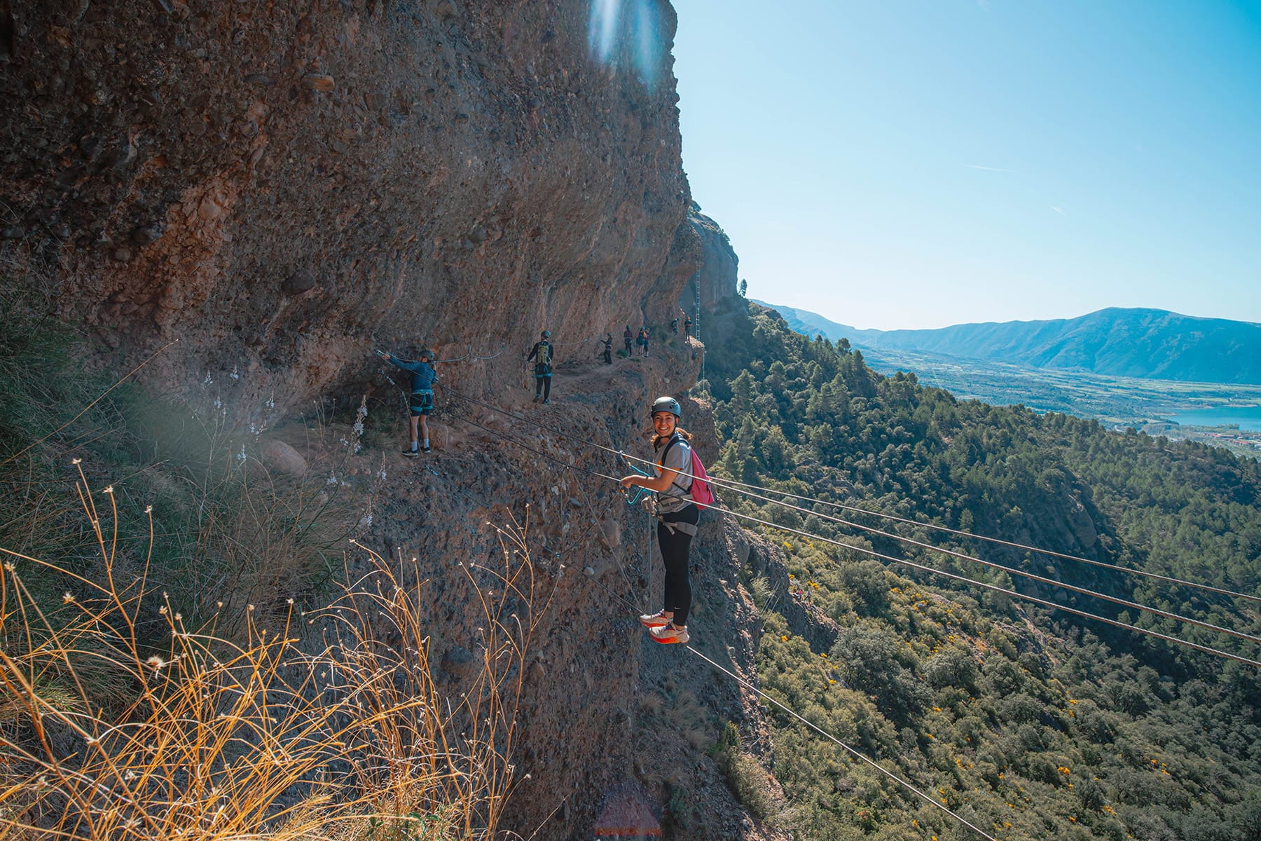 Climber crossing the exposed cable bridge on the Vía Ferrata Roca Foradada, with other participants along the rock wall and panoramic views near La Pobla de Segur