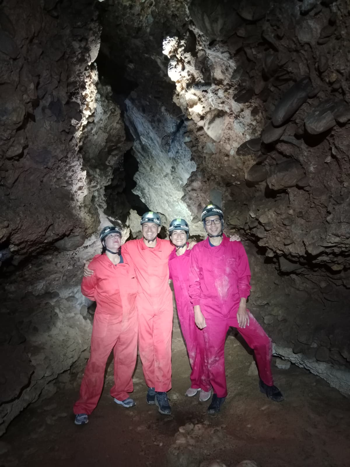 Group of four cavers posing together inside Cueva Forat la Bou wearing protective suits, helmets and headlamps