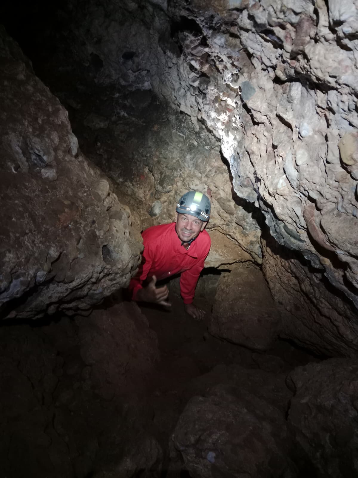 Caver climbing through a narrow rocky passage inside Cueva Forat la Bou wearing a red suit, helmet and headlamp