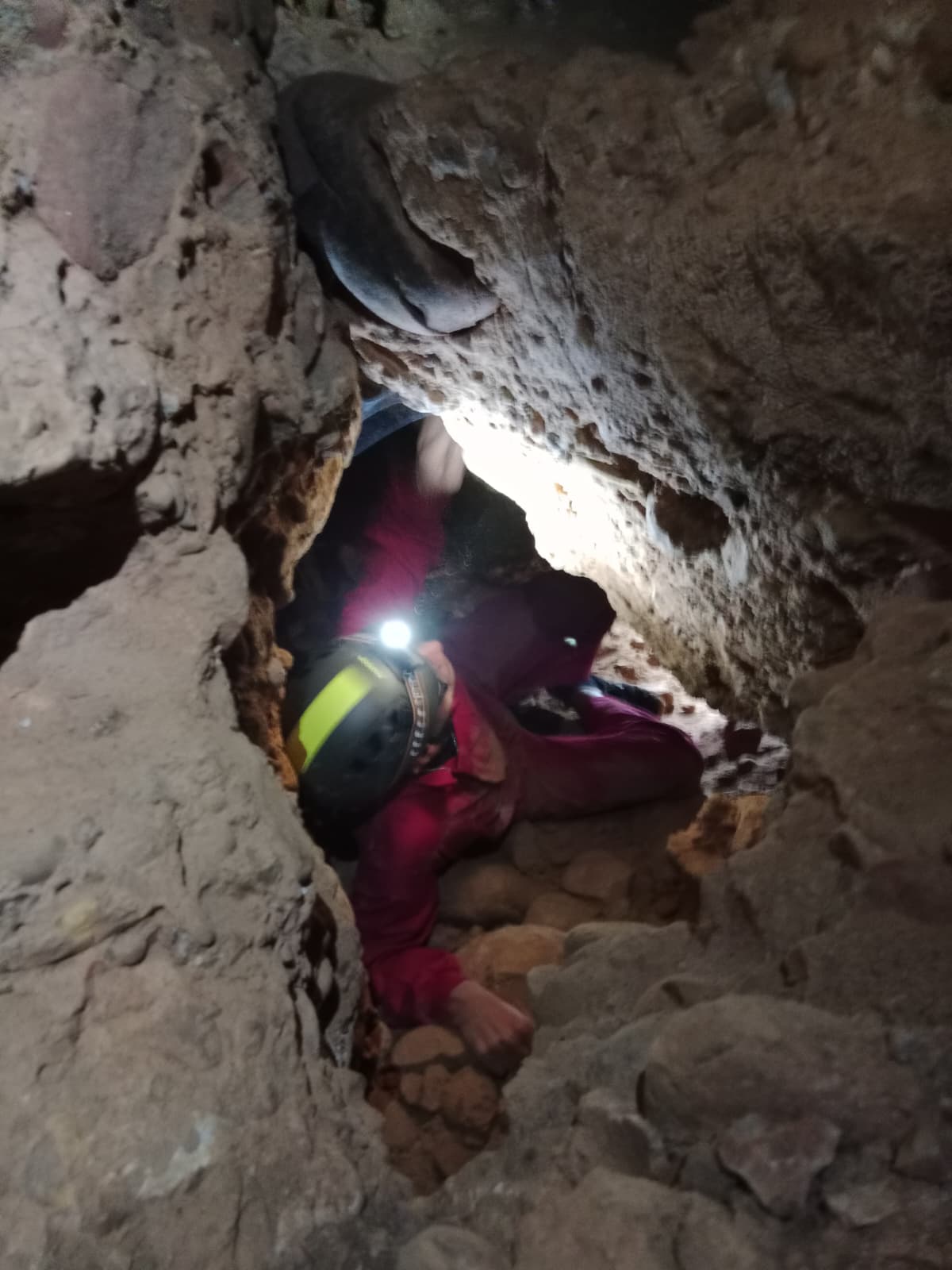 Caver crawling through an extremely narrow rocky passage inside Cueva Forat la Bou wearing a helmet and headlamp