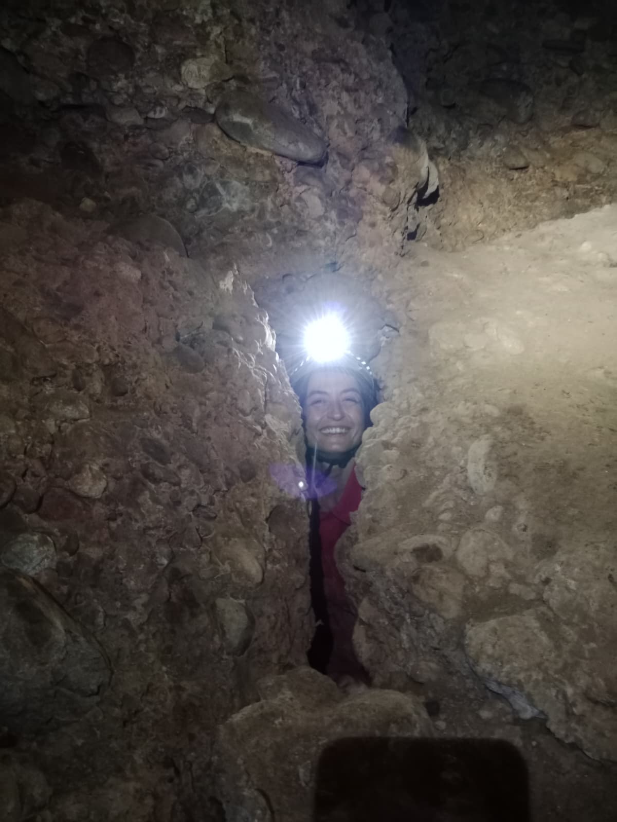 Smiling caver squeezing through a very narrow rocky passage inside Cueva Forat la Bou wearing a helmet and headlamp