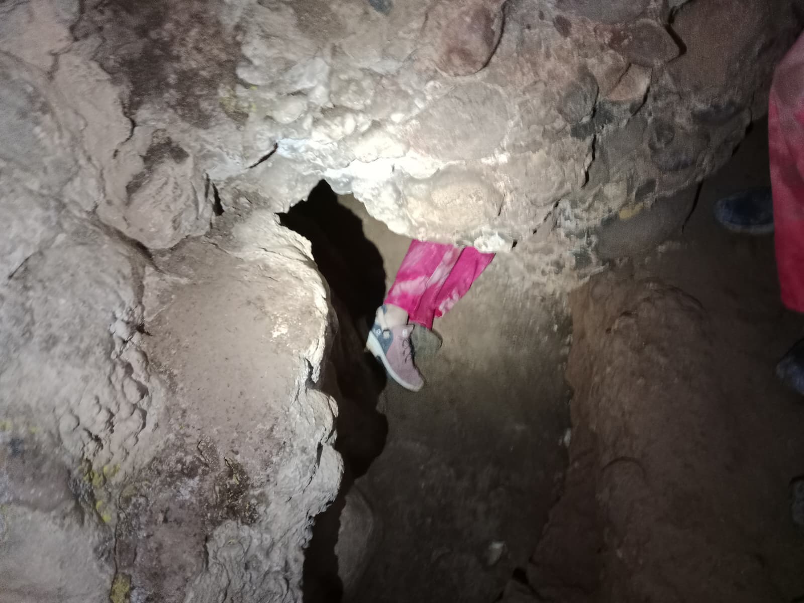 Caver squeezing feet first through a narrow rocky opening inside Cueva Forat la Bou wearing a protective suit