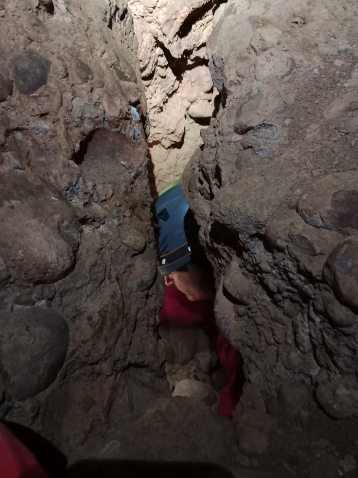 Caver squeezing through an extremely narrow rocky passage inside Cueva Forat la Bou wearing a helmet and protective suit