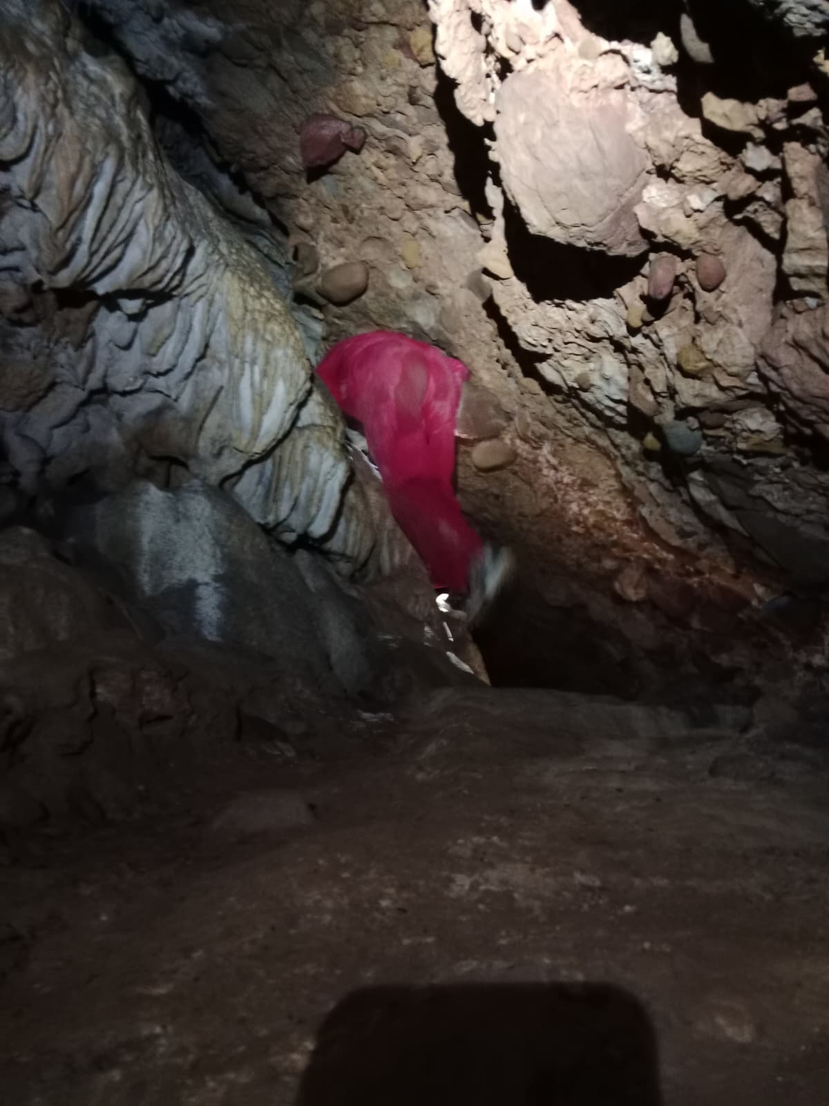 Caver descending through a steep rocky passage inside Cueva Forat la Bou wearing a red protective suit and helmet