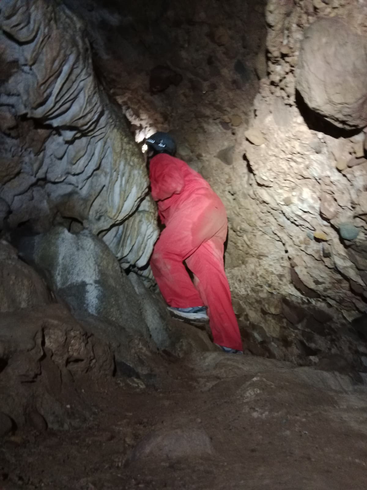 Caver climbing through a rocky passage inside Cueva Forat la Bou, wearing a red protective suit and helmet among sculpted cave formations