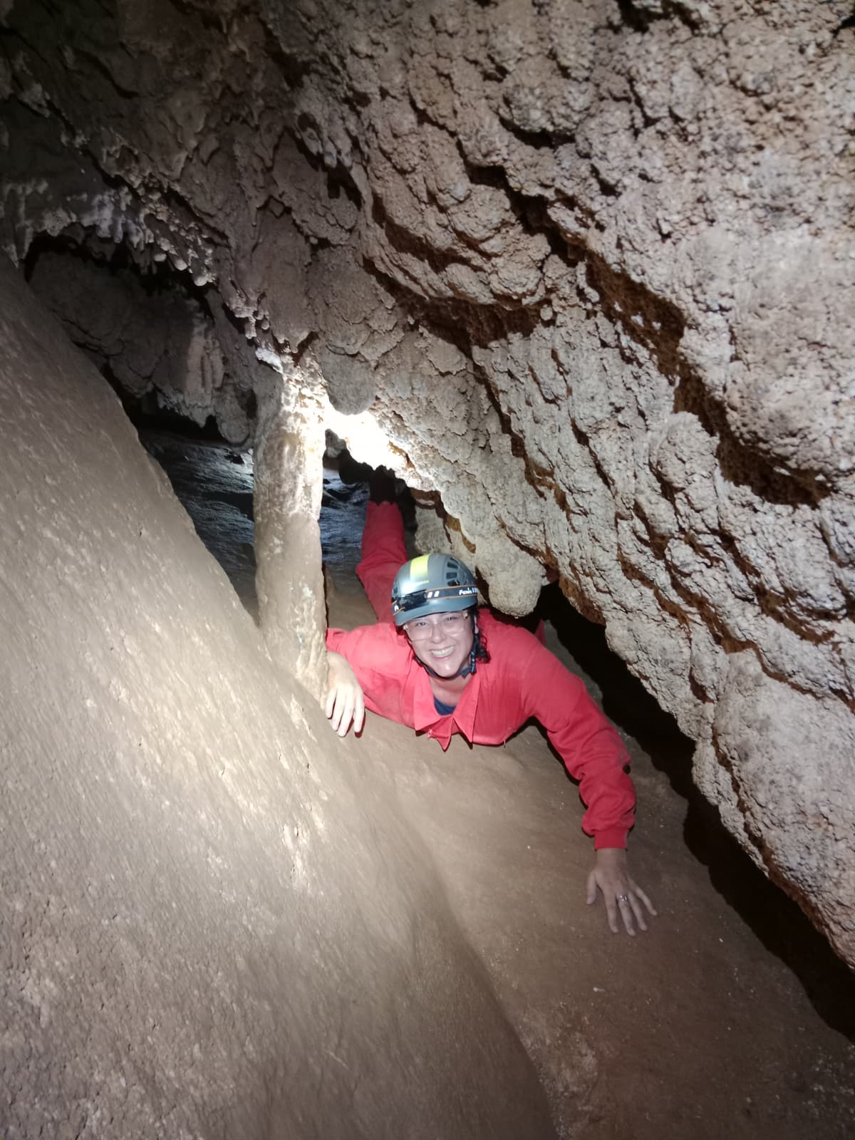 Smiling caver crawling through a narrow passage inside Cueva Forat la Bou wearing a protective suit, helmet and headlamp